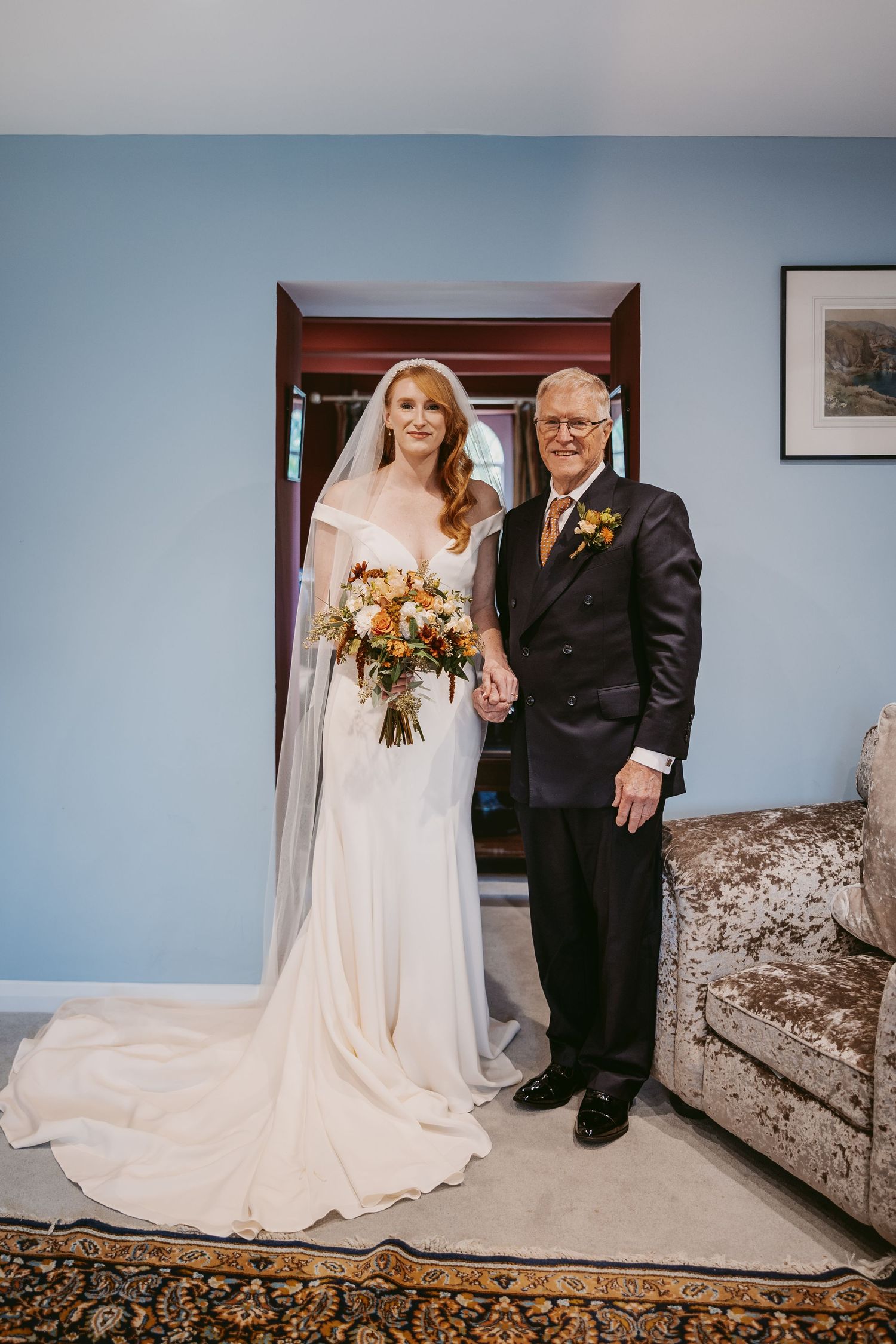 Bride in an elegant white gown and veil holds her bouquet and stands hand in hand with her father in a blue-walled room.
