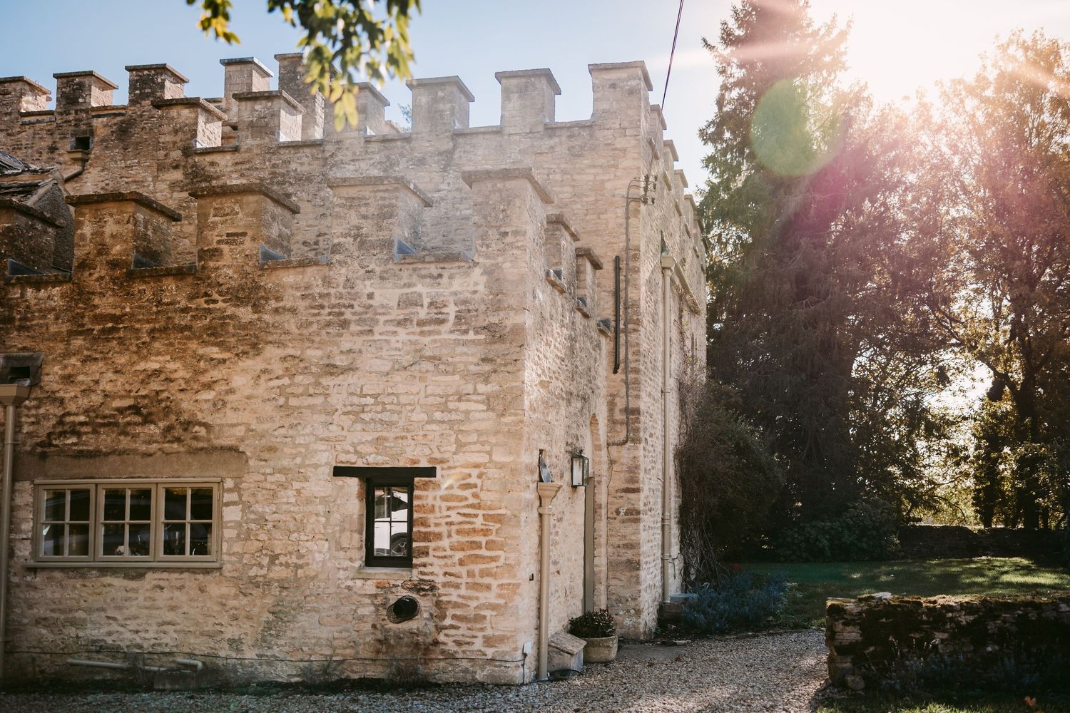 Sunlight streams over a crenellated stone castle wedding venue with tall trees and a small window in the golden light.