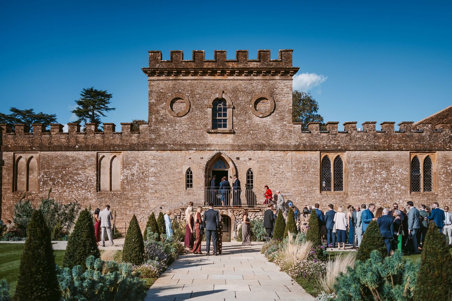 Wedding guests mingle on a gravel path lined with topiary in front of a historic castle-style stone building with battlements.