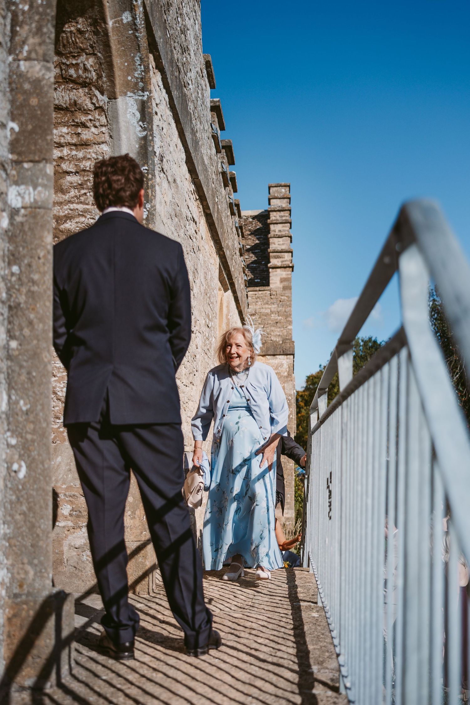 A man in a dark suit stands on a sunlit stone walkway as an elderly woman in a blue dress approaches beside a historic wall.