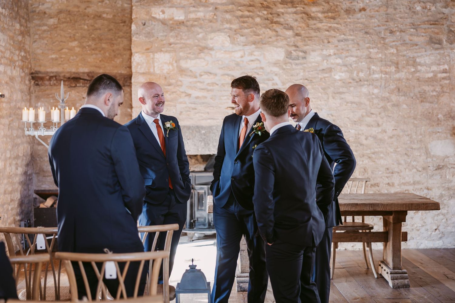 Groom and groomsmen in dark suits chat together in a rustic stone-walled room with wooden tables and chairs before the wedding.