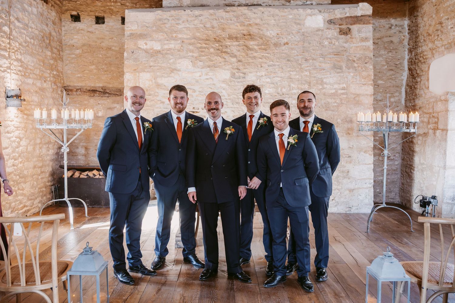 Six groomsmen in navy suits with orange ties and buttonholes pose together in a rustic stone room with candle holders and lanterns.