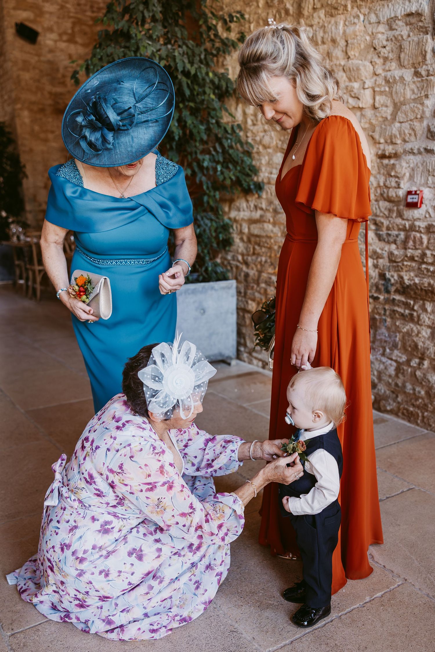 Two women in blue and floral outfits and a bridesmaid in rust orange greet a toddler in a waistcoat at a wedding.