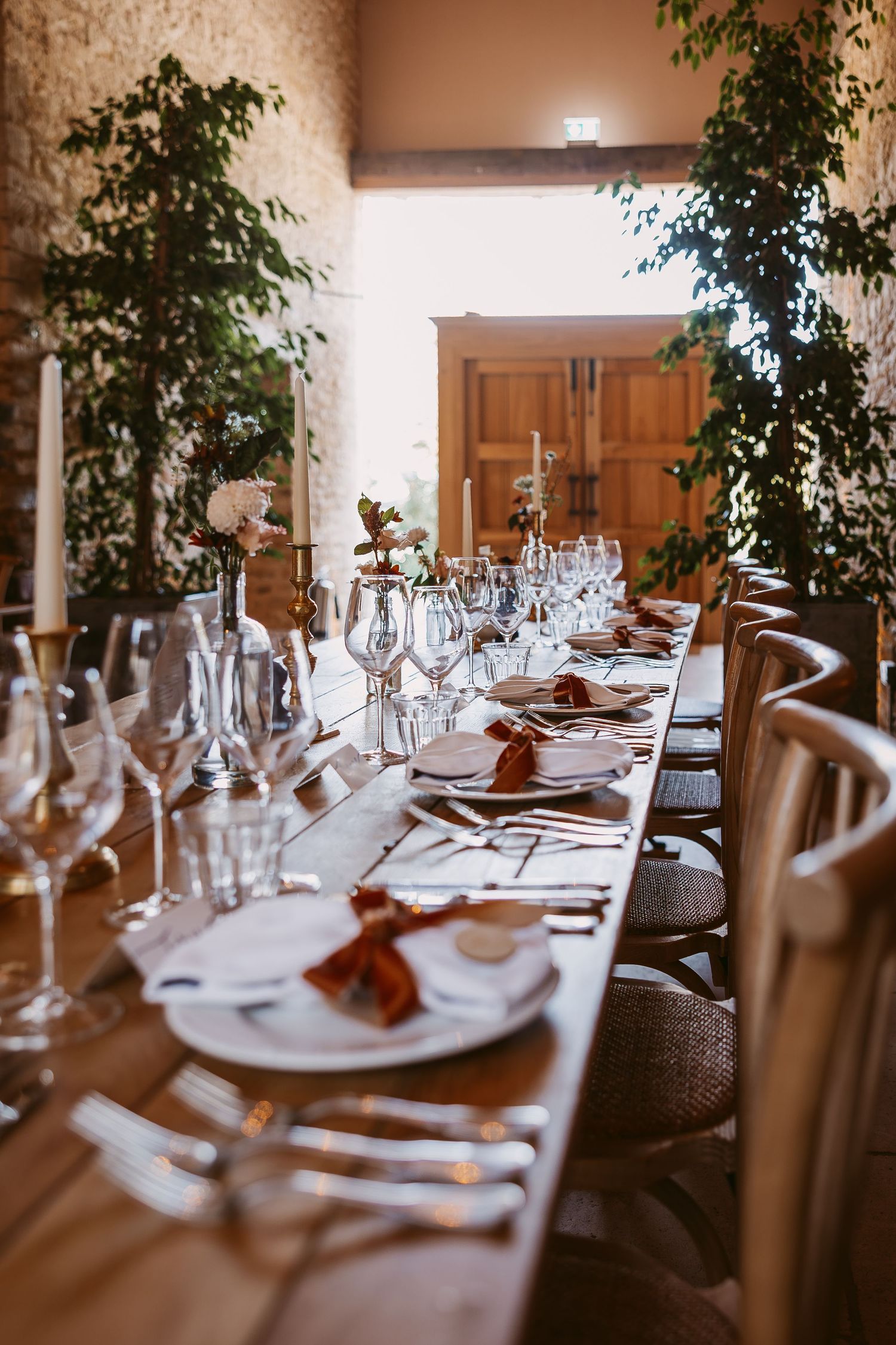 Long rustic wooden wedding table set with glassware, plates, rust napkins and candles in a stone barn venue.