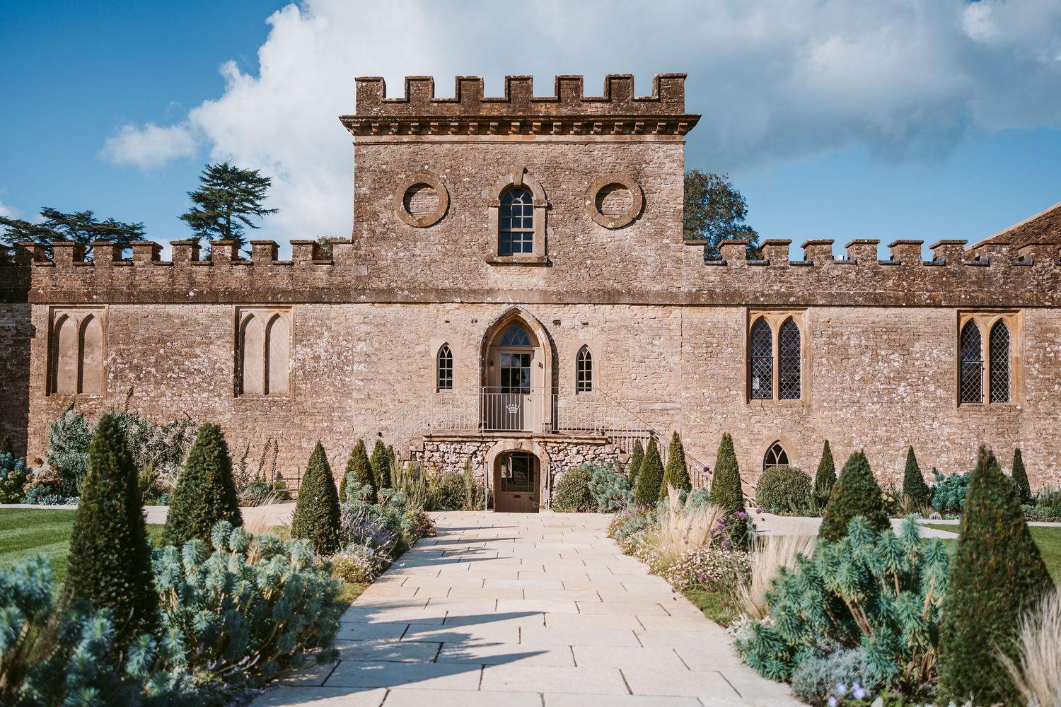 Crenellated stone castle wedding venue with a gravel path and clipped topiary shrubs under a blue sky with clouds.