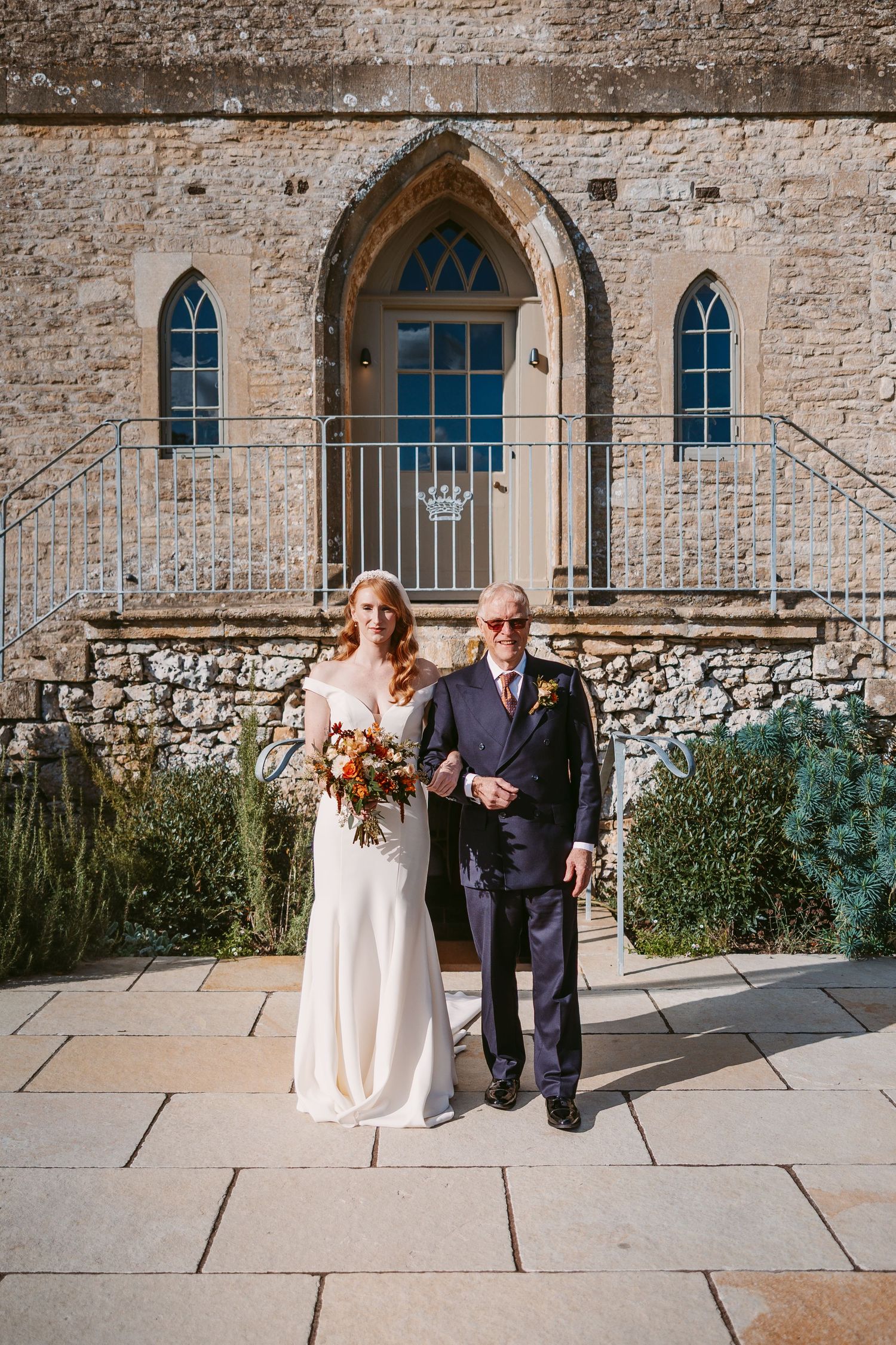 Bride in an off-shoulder gown holds an autumn bouquet while standing arm in arm with her father outside a stone chapel.
