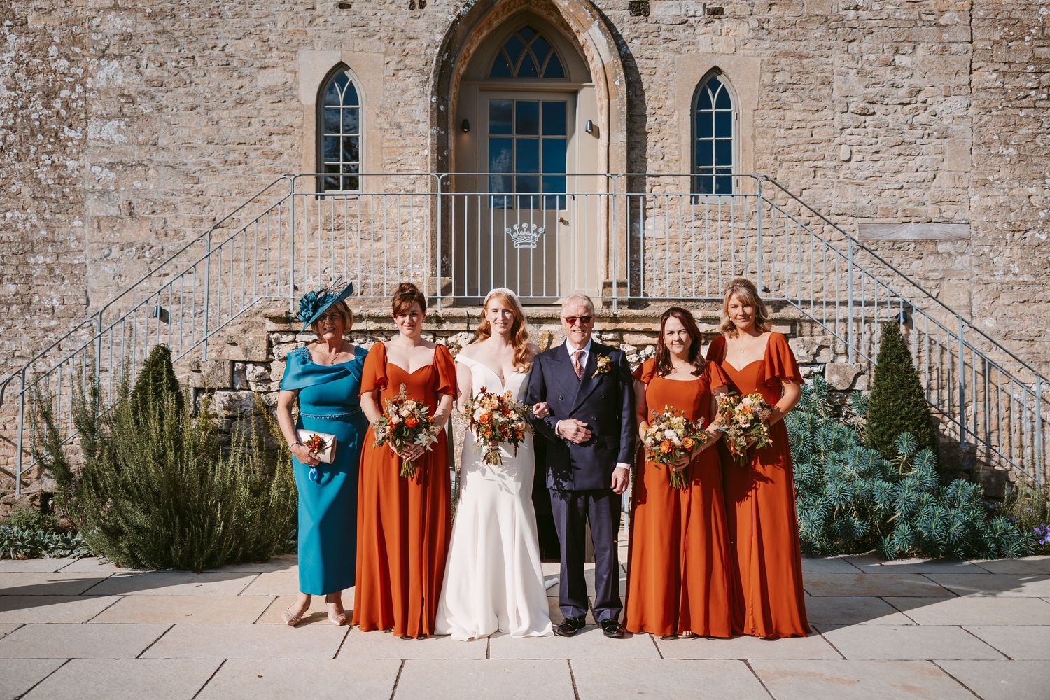 Bride, groom and four women in blue and rust orange dresses pose with bouquets in front of a stone church entrance.