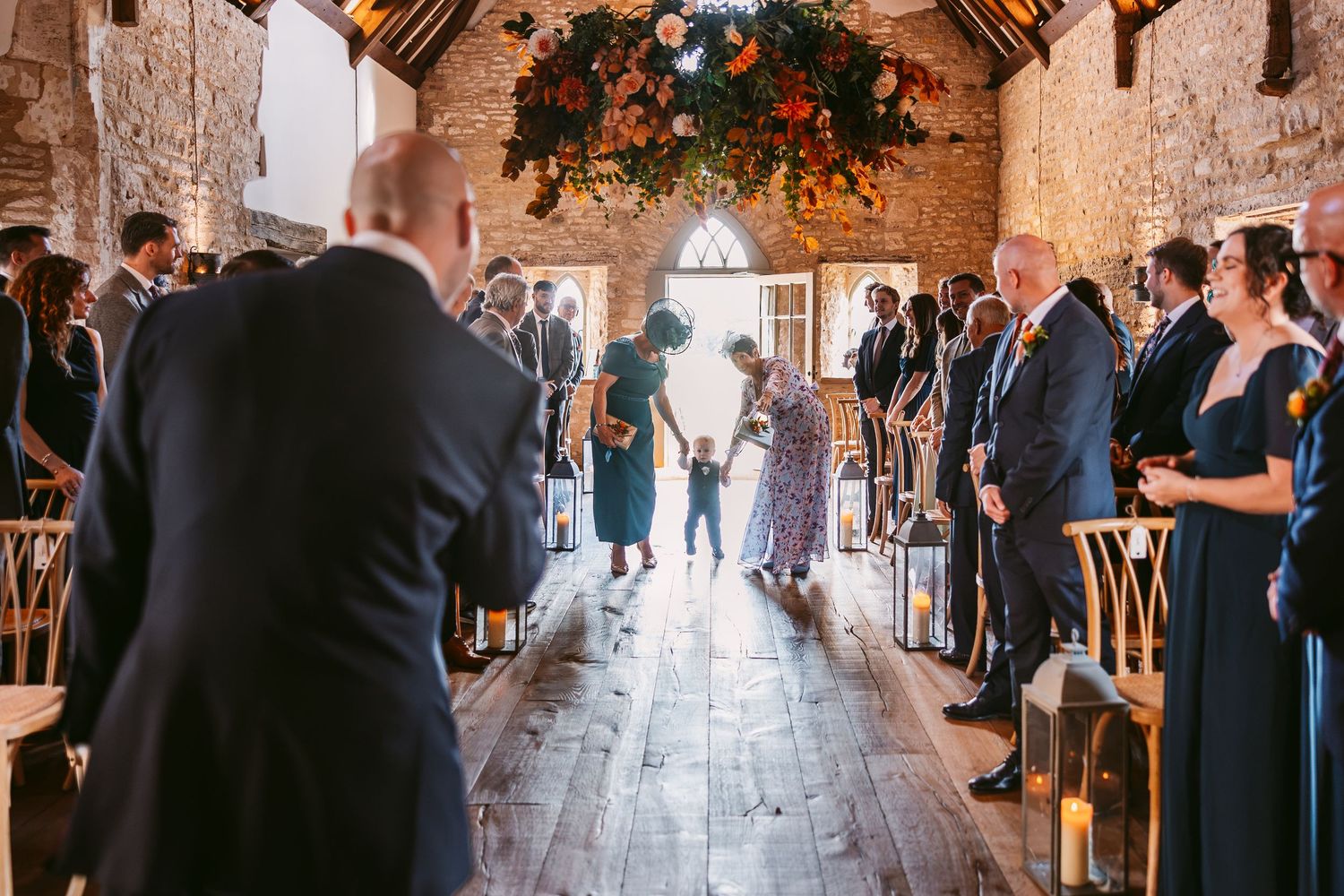 Wedding guests stand along a rustic barn aisle as a flower girl walks toward the altar beneath a hanging floral chandelier.