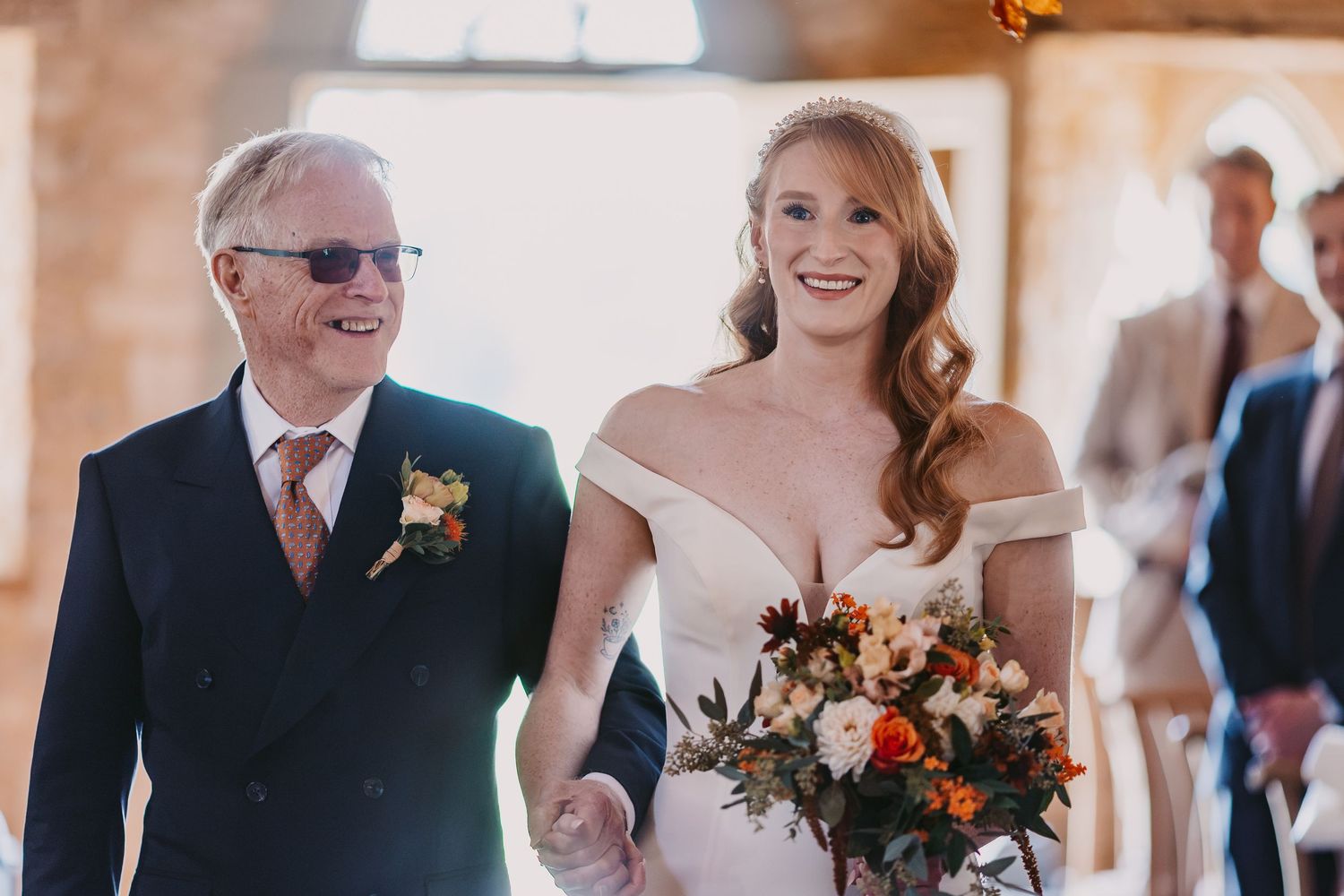 Smiling bride with an orange and white bouquet walks down the aisle holding the hand of her father in a navy suit.