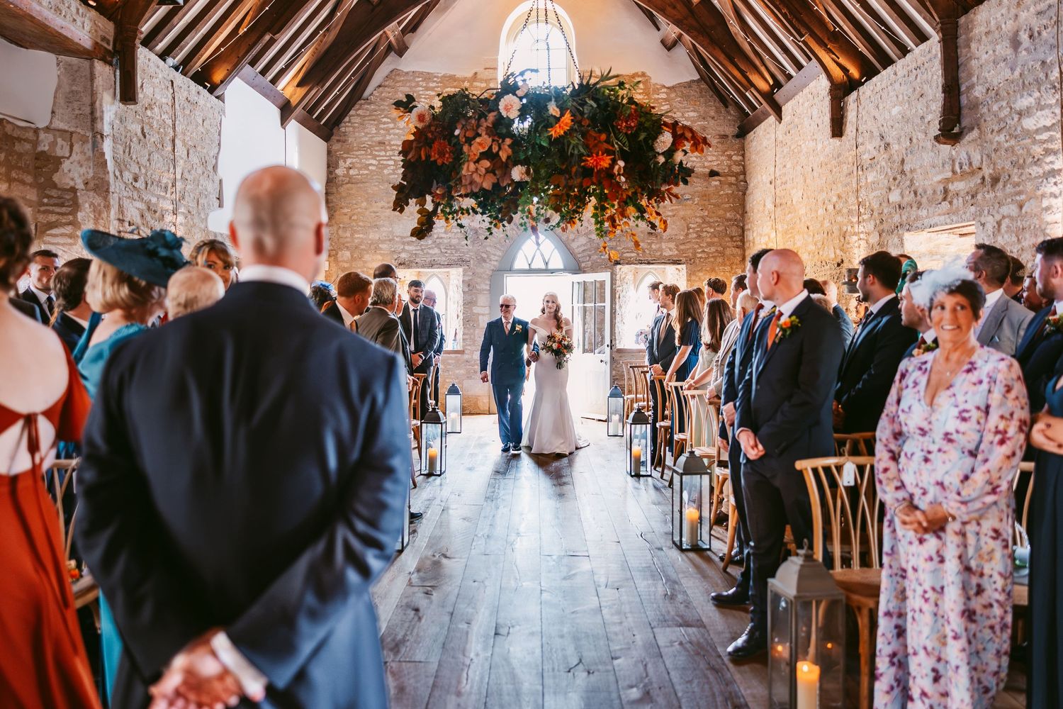 Bride in white gown walks down the aisle of a rustic stone barn wedding venue beneath a large autumn floral installation.
