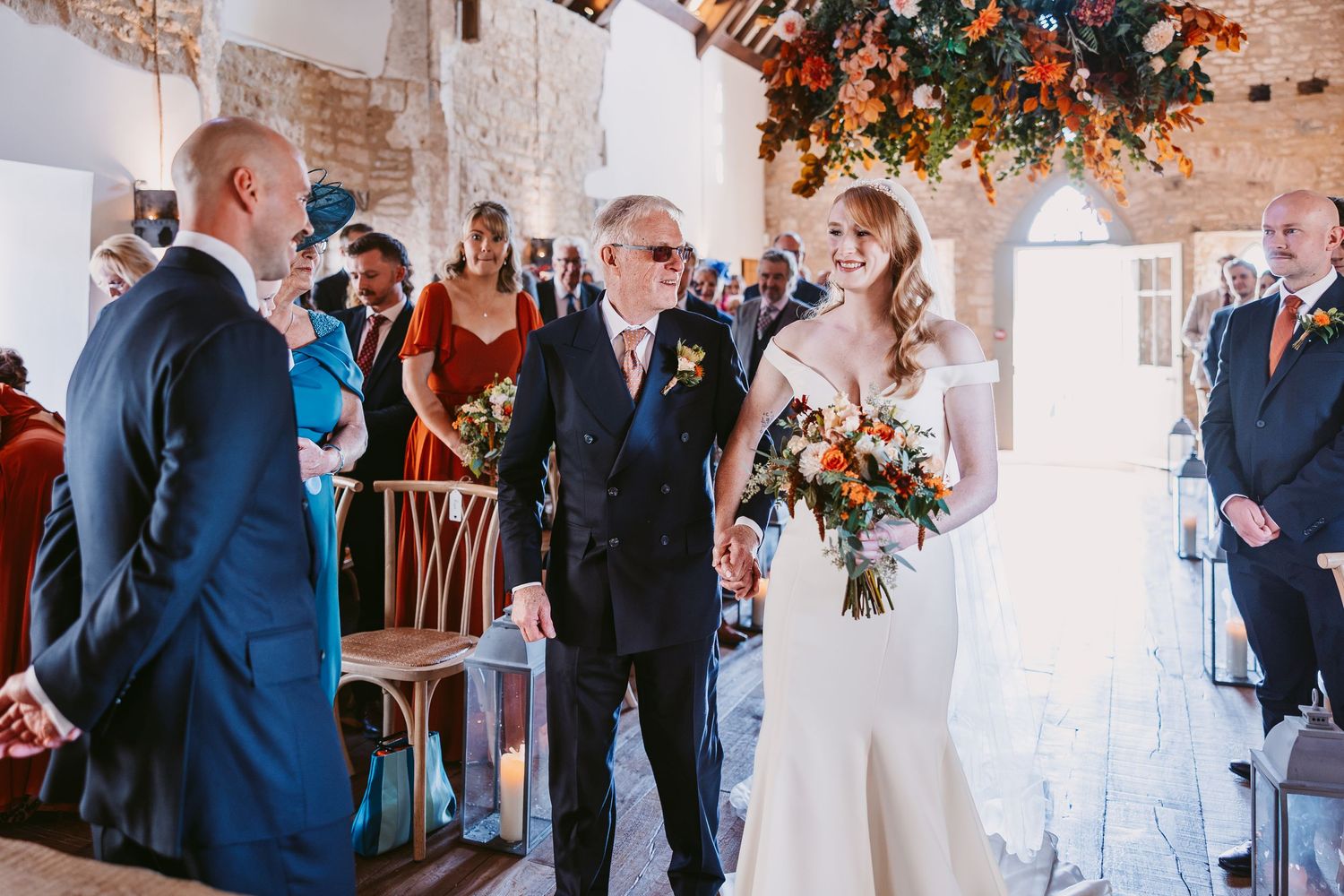 Smiling bride with an orange bouquet walks down the aisle on her father's arm past seated wedding guests.