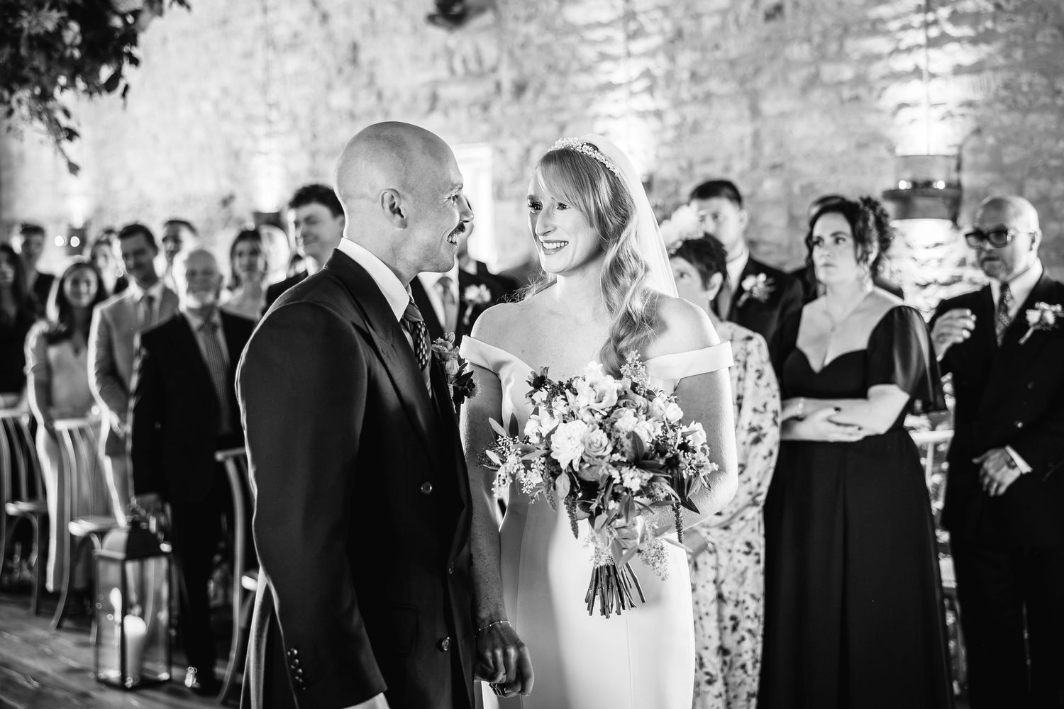 Black and white photo of a smiling groom and bride holding a bouquet as wedding guests look on during the ceremony.