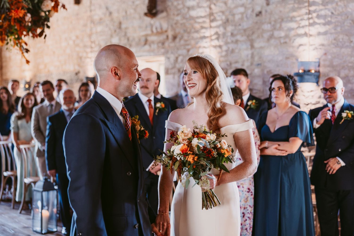 Bride in a white gown holding an orange autumn bouquet shares a smile with her groom during their wedding ceremony.