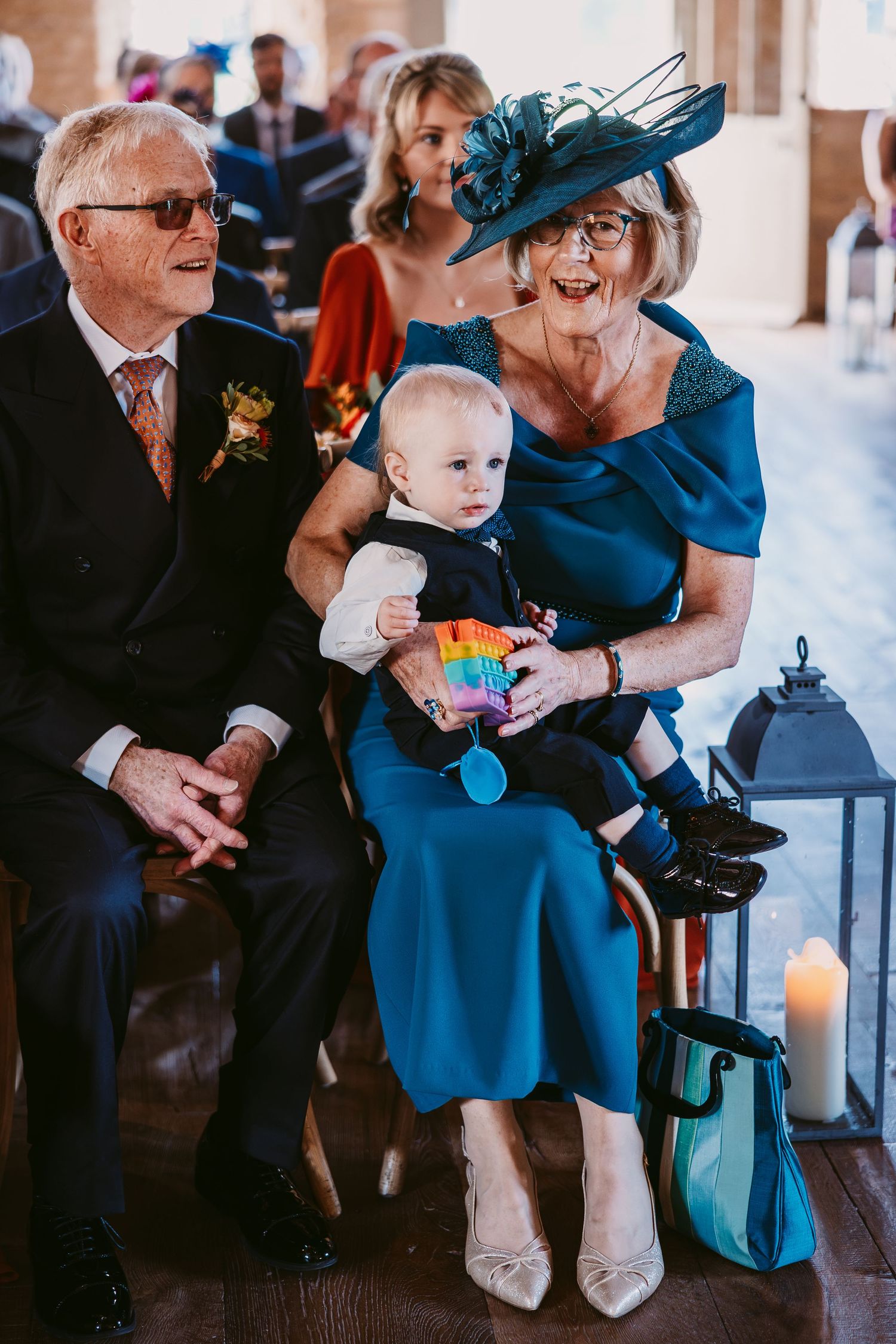 Elderly man and woman in a teal outfit and fascinator hold a smiling toddler with a rainbow toy during a wedding ceremony.