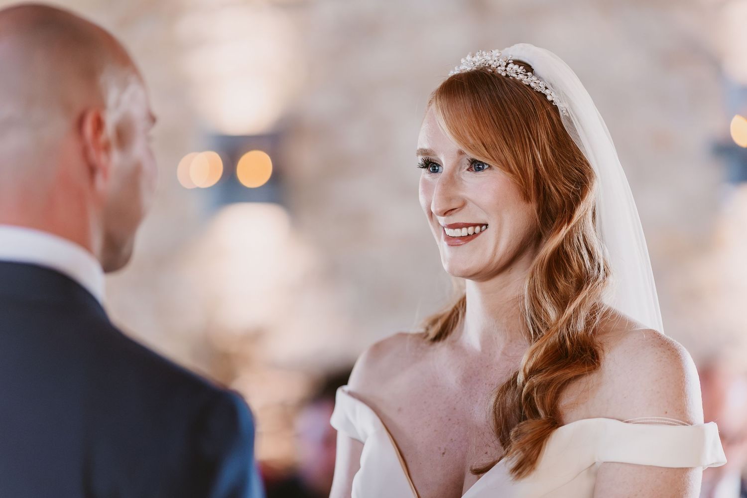 A smiling red-haired bride in an off-shoulder dress and tiara veil gazes at her groom during the wedding ceremony.
