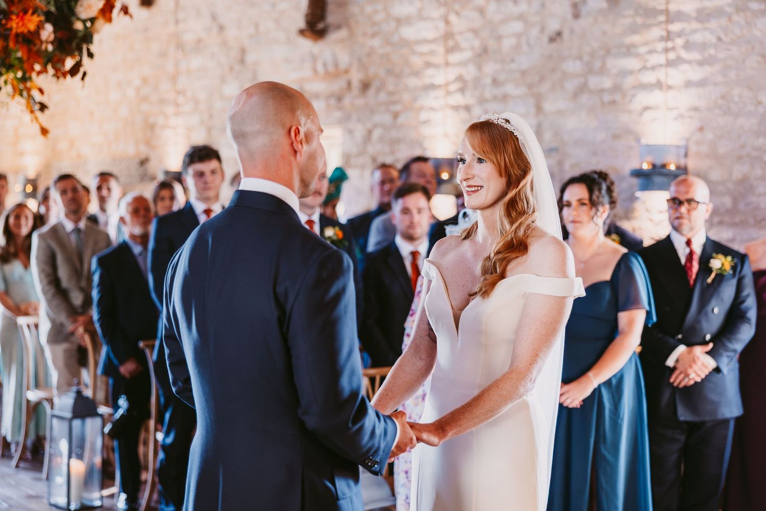 A bride and groom hold hands facing each other at the altar while wedding guests watch in a rustic stone venue.