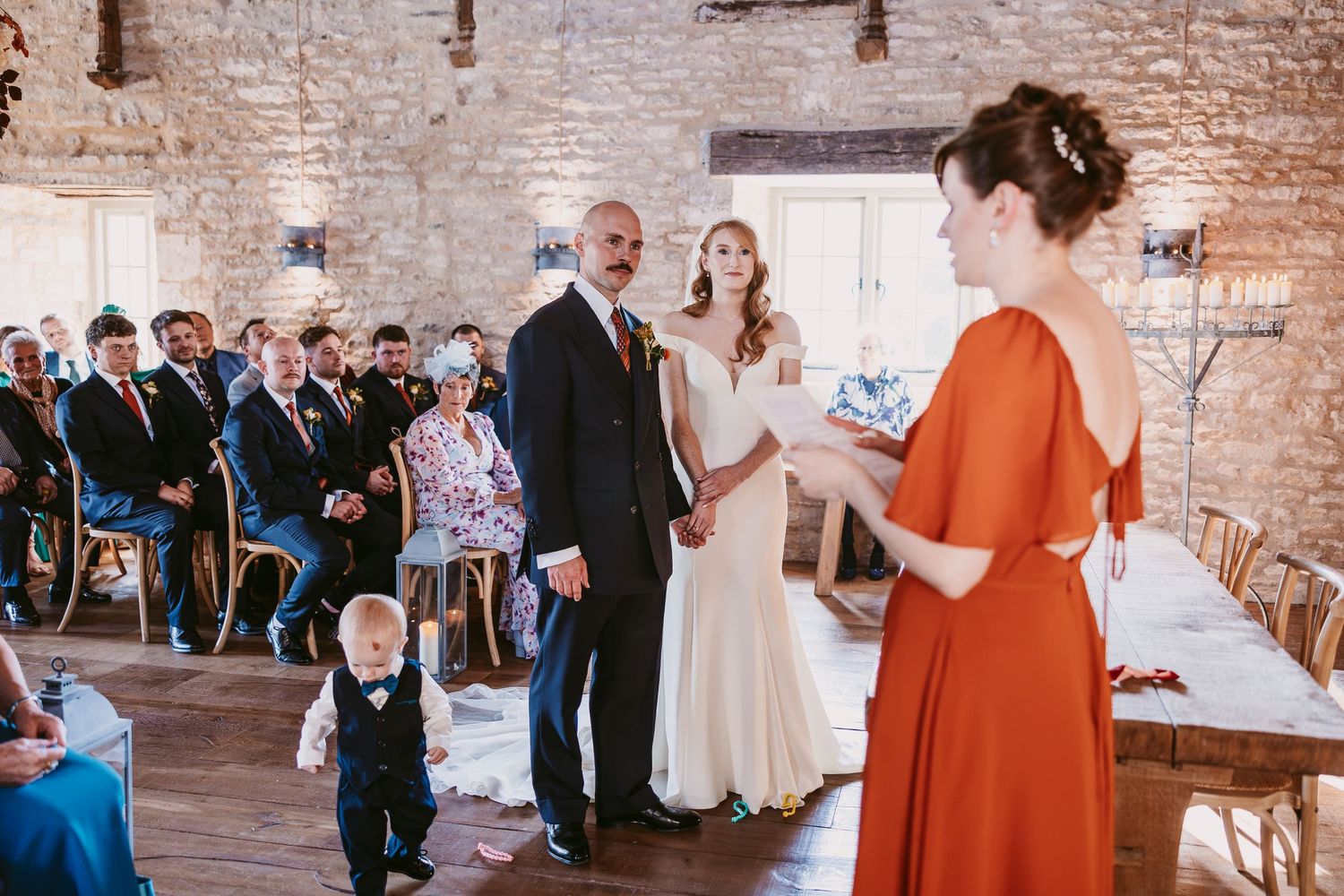 Woman in an orange dress reads to a bride and groom during a barn wedding ceremony while a toddler in a waistcoat watches.