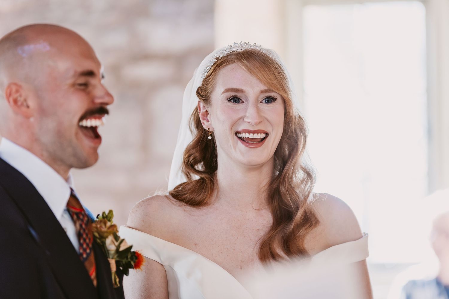 Laughing groom and smiling bride in a tiara and veil share a joyful moment during their wedding ceremony.
