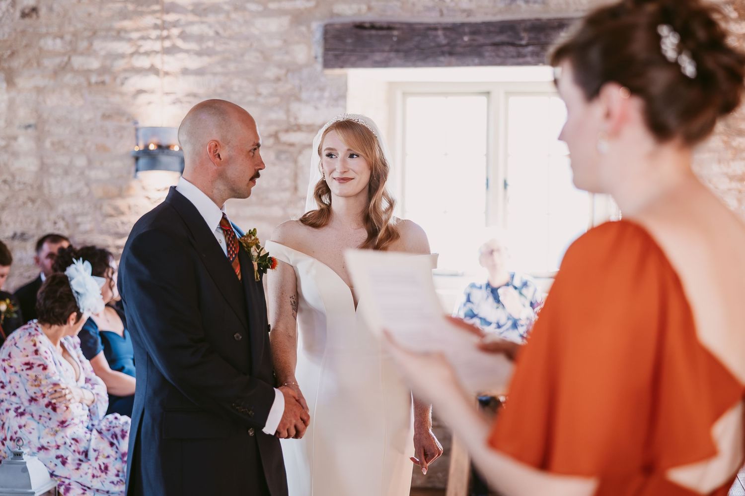 A bride and groom hold hands during their wedding ceremony in a rustic stone venue while a woman in orange reads aloud.