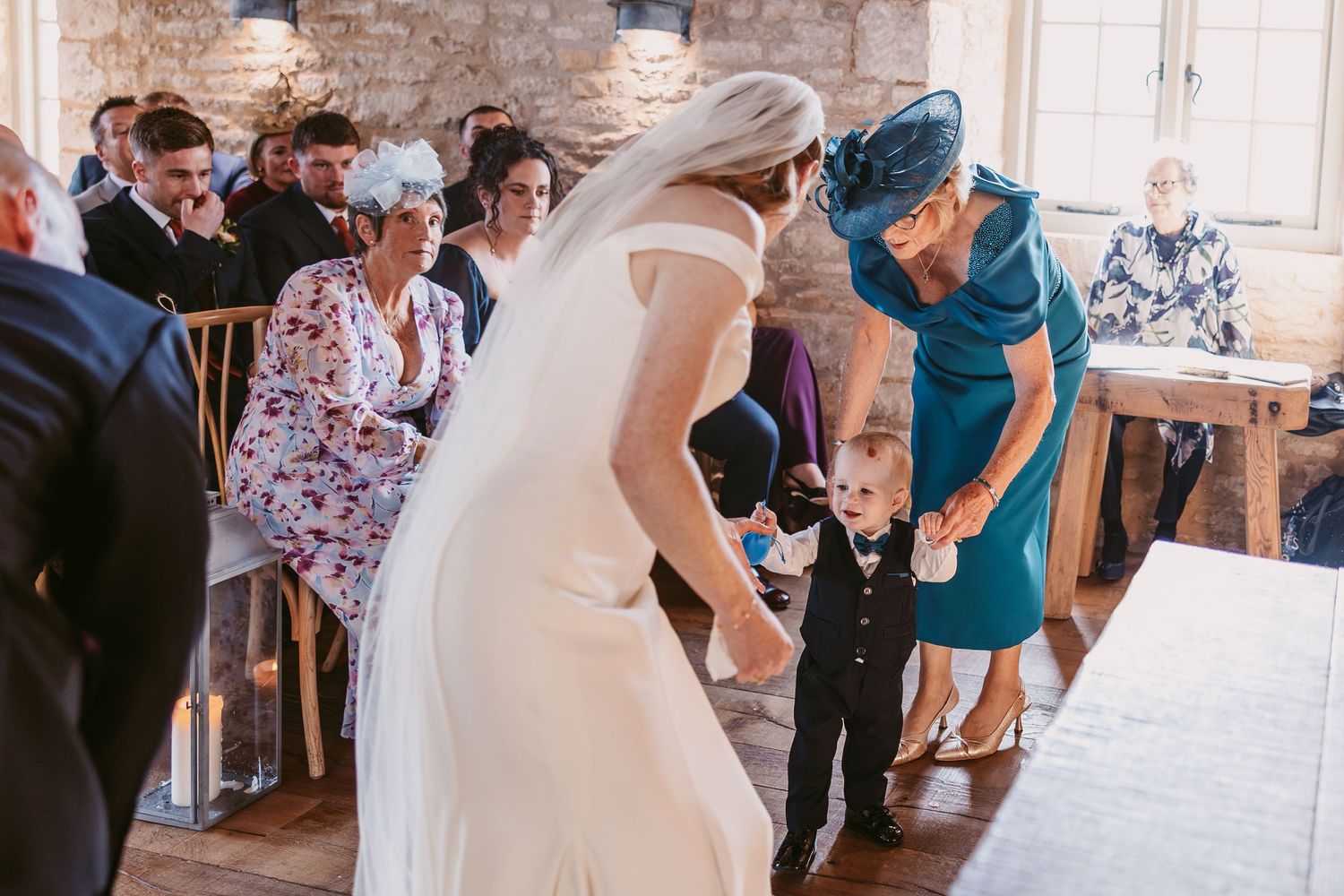 Bride in a flowing veil and a guest in a teal dress greet a toddler in a black suit during a wedding ceremony.