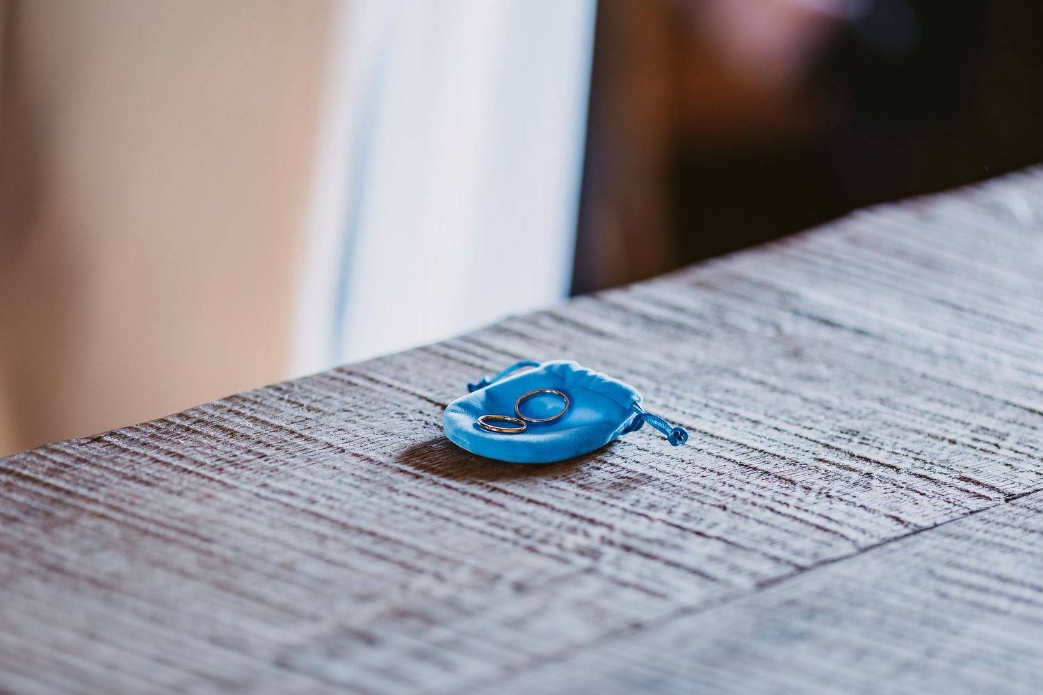 A small blue ring dish holding a wedding ring rests on a rustic weathered wooden surface with soft blurred light behind it.