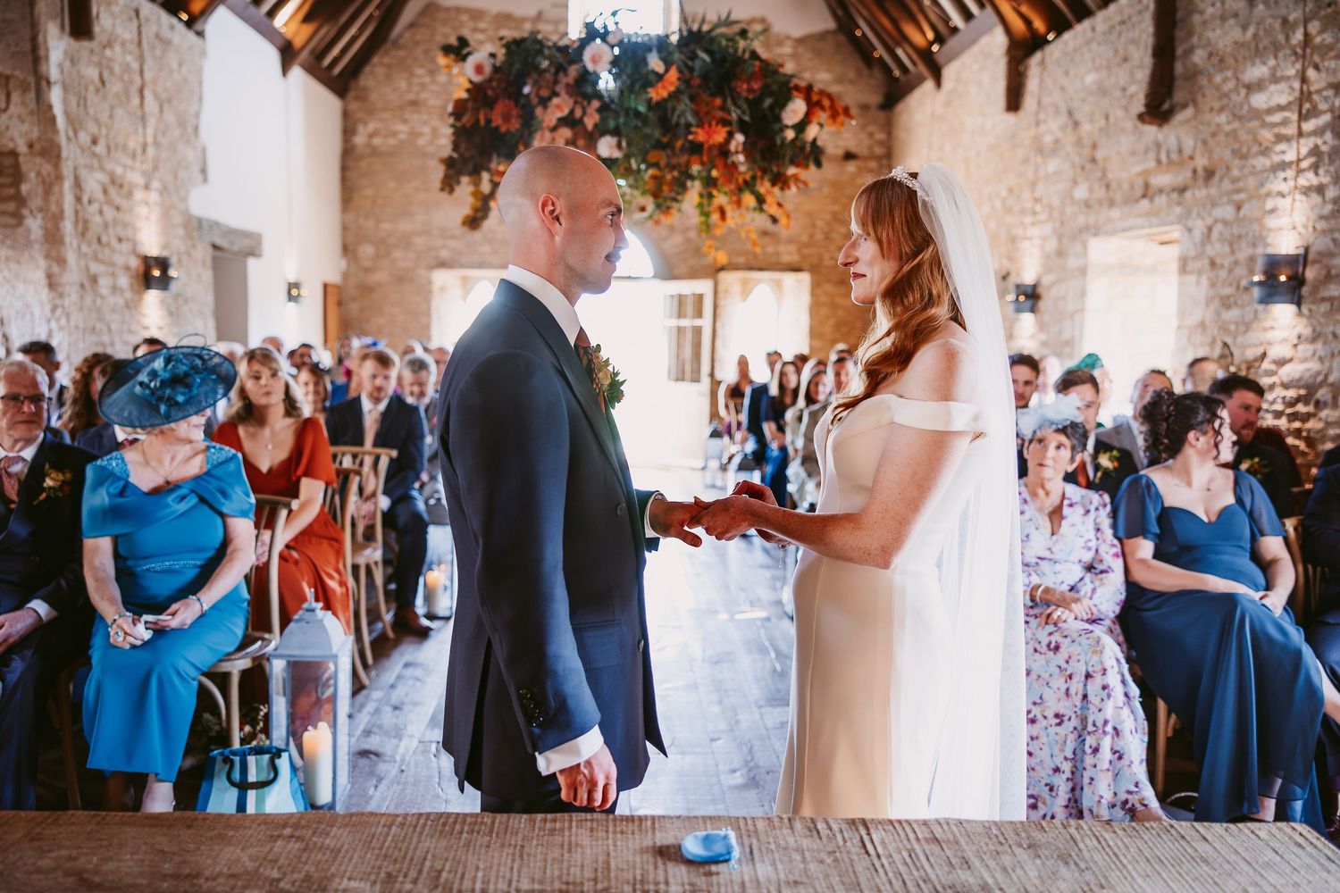 Bride and groom hold hands at the registry table during their rustic barn wedding ceremony as seated guests watch.