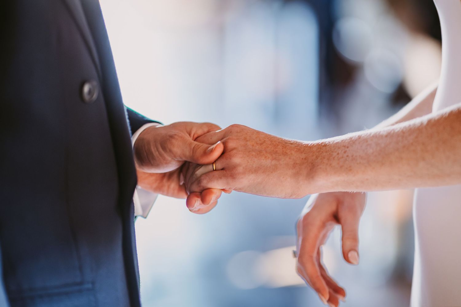 Close-up of a bride and groom clasping hands during their wedding ceremony, with a wedding ring visible on the bride's finger.