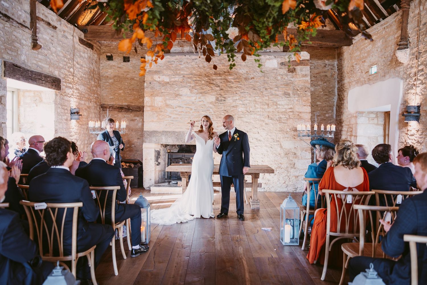 Bride and groom exchange vows during a wedding ceremony in a rustic barn with exposed beams, brick walls and hanging autumn foliage.