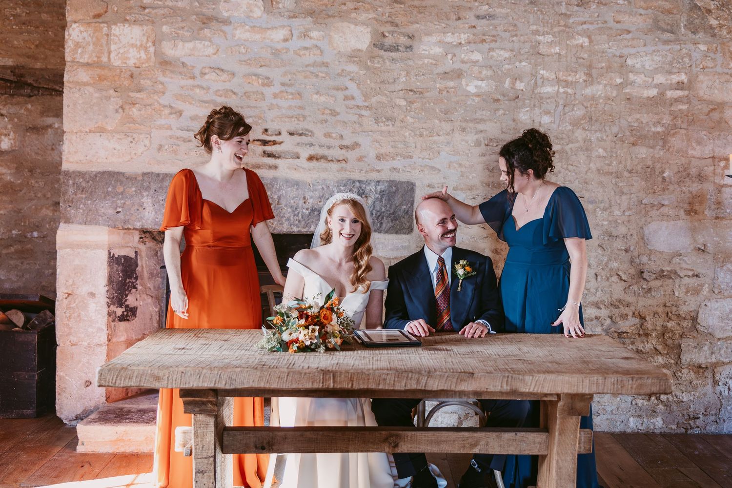 A bride and groom sit at a rustic wooden table signing the wedding register while two bridesmaids in orange and teal stand beside them.