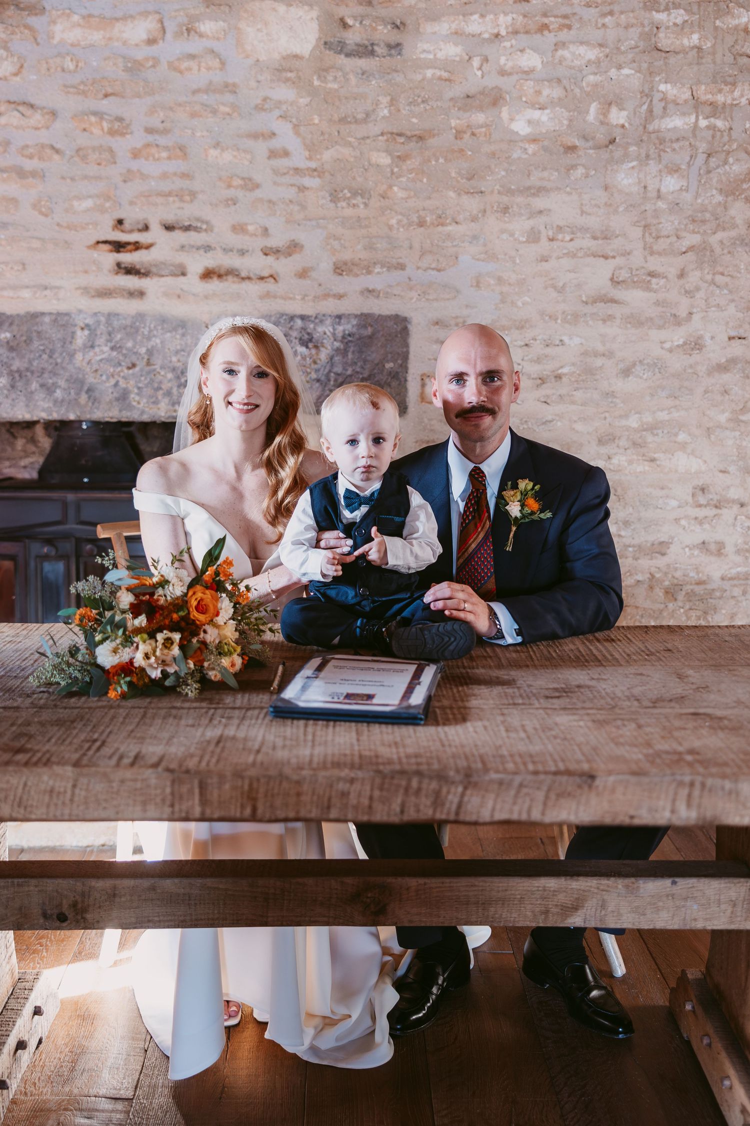 Bride in a white gown and groom with a waxed mustache sit at a wooden table with their toddler son and an orange floral bouquet.