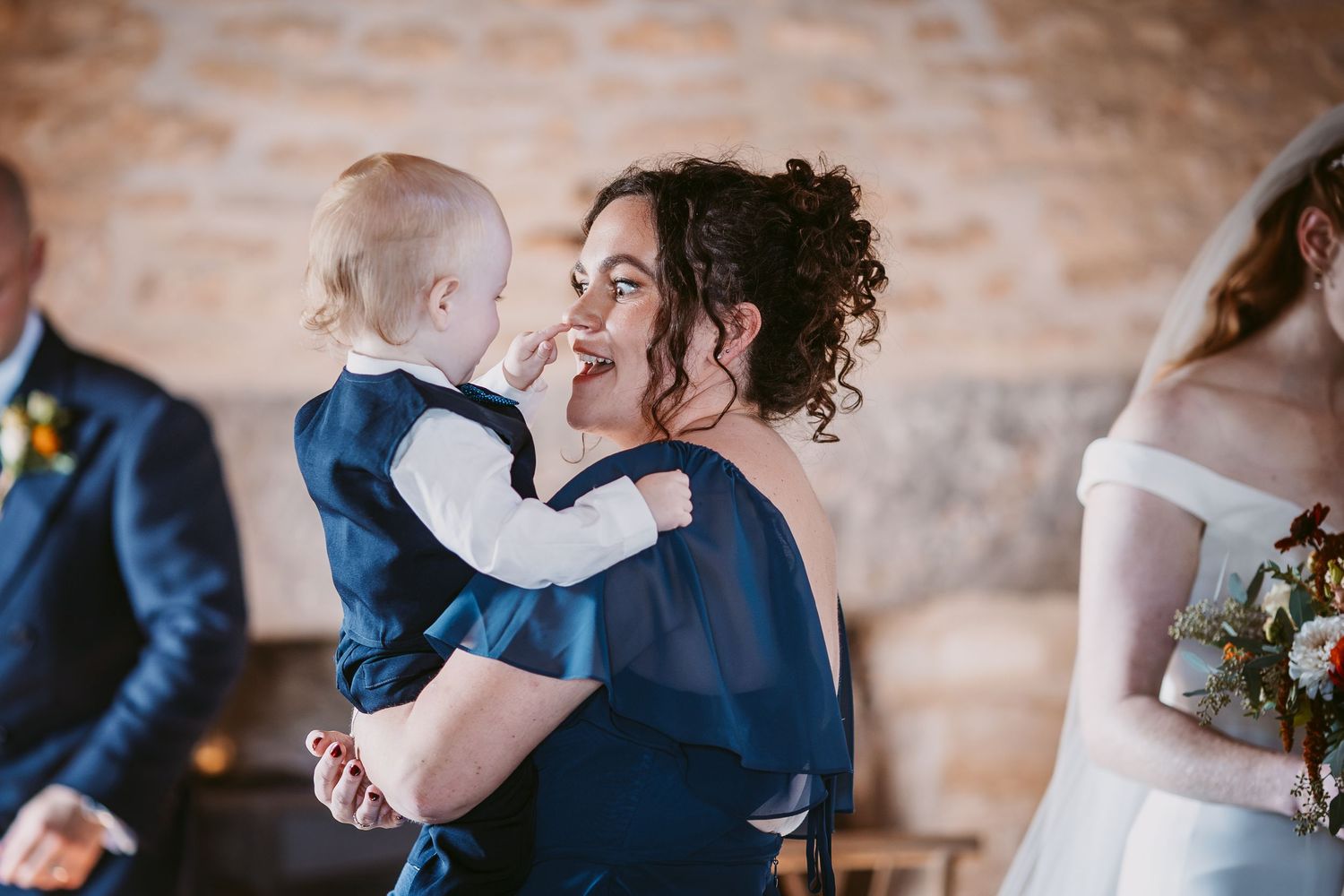 Smiling woman in a navy dress holds a baby in a matching outfit at a wedding while a bride with a floral bouquet stands beside her.