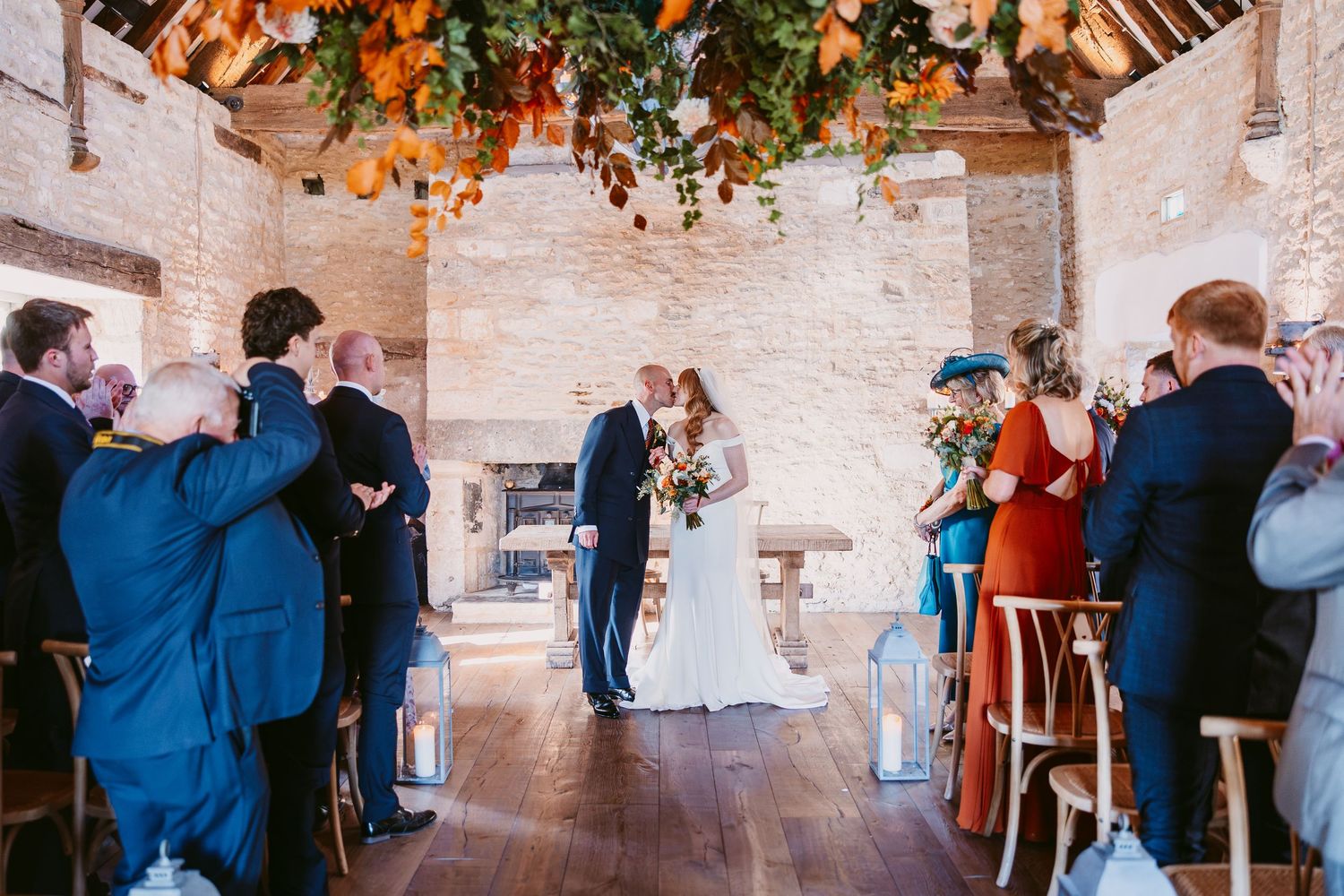 Bride and groom share a moment at the altar of a rustic barn wedding ceremony beneath hanging autumn floral decor as guests watch.