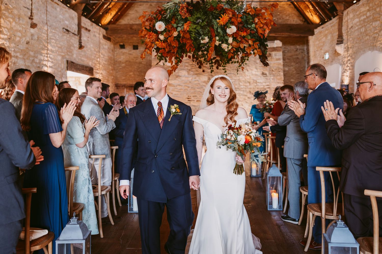 Smiling bride and groom walk down the aisle as wedding guests cheer and applaud in a rustic barn venue with a hanging floral installation.