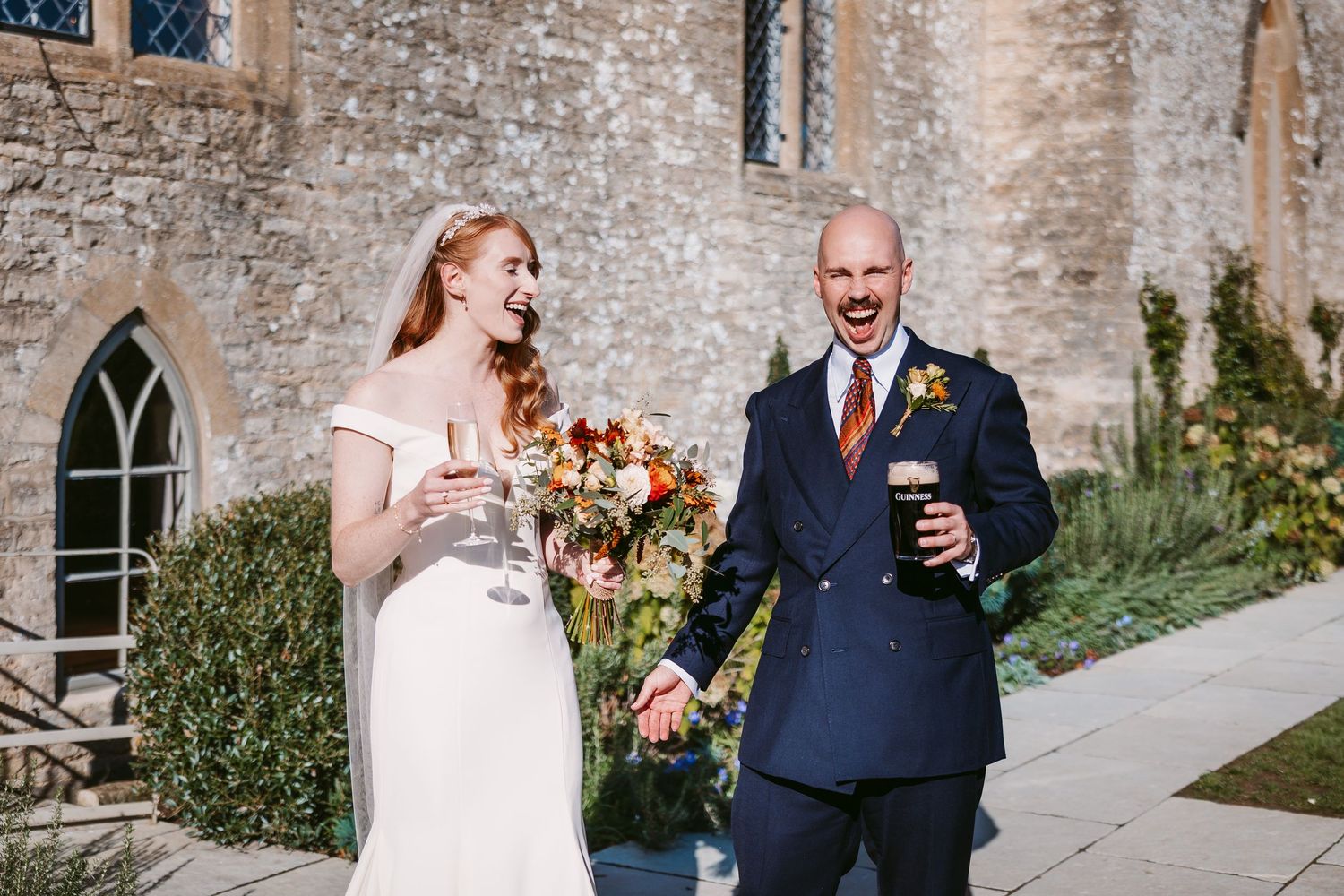 Laughing bride in a white gown holding a bouquet and champagne flute beside a groom in a navy suit holding a pint outside a stone building.