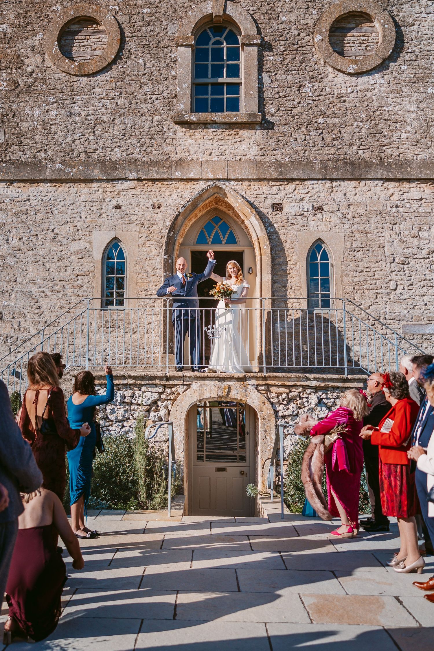 Bride and groom raise their arms in celebration from a stone church doorway as wedding guests cheer below on a sunlit terrace.