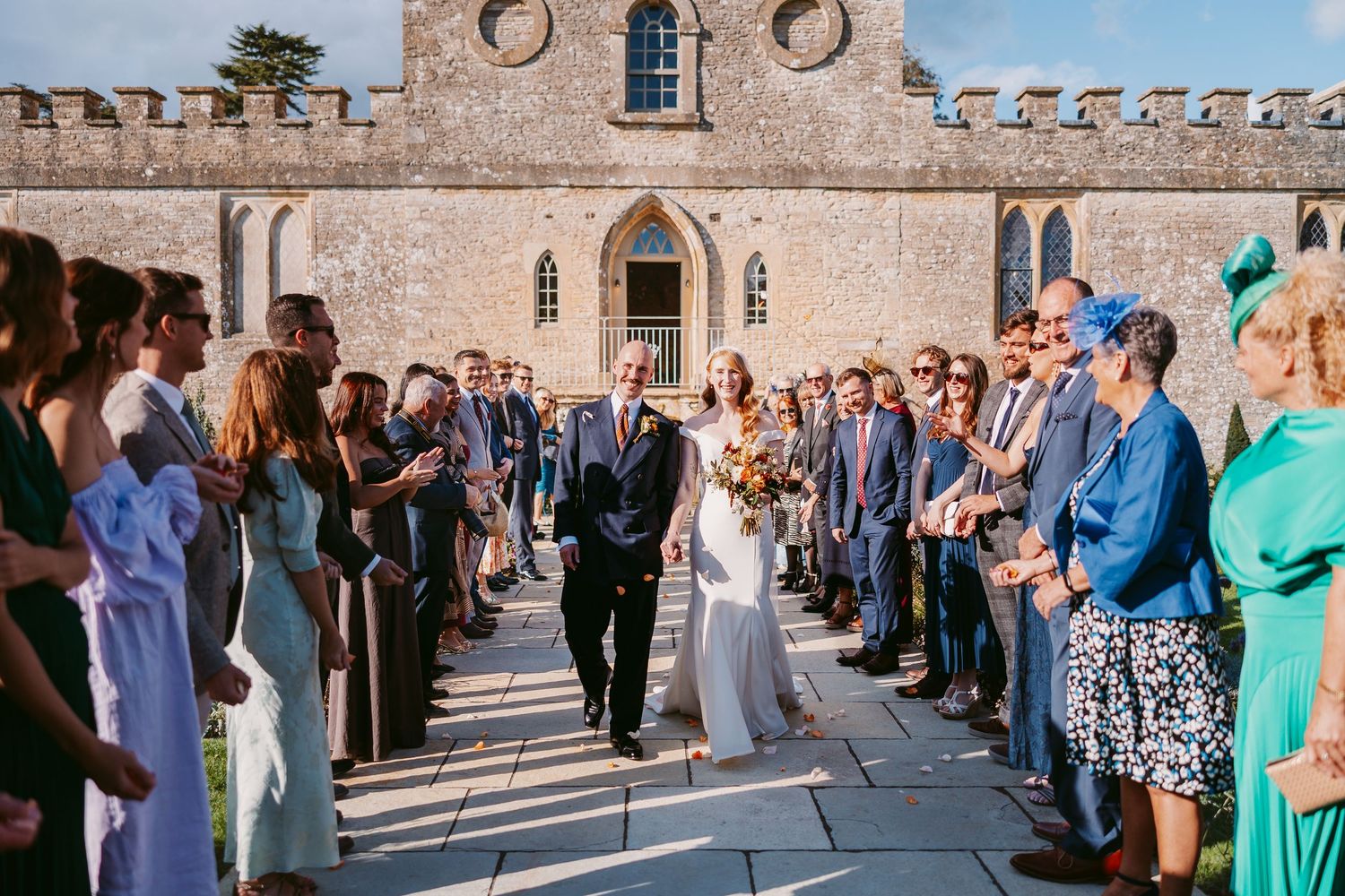Bride in a white gown and groom in a dark suit walk down a stone path past cheering wedding guests outside a historic castle venue.