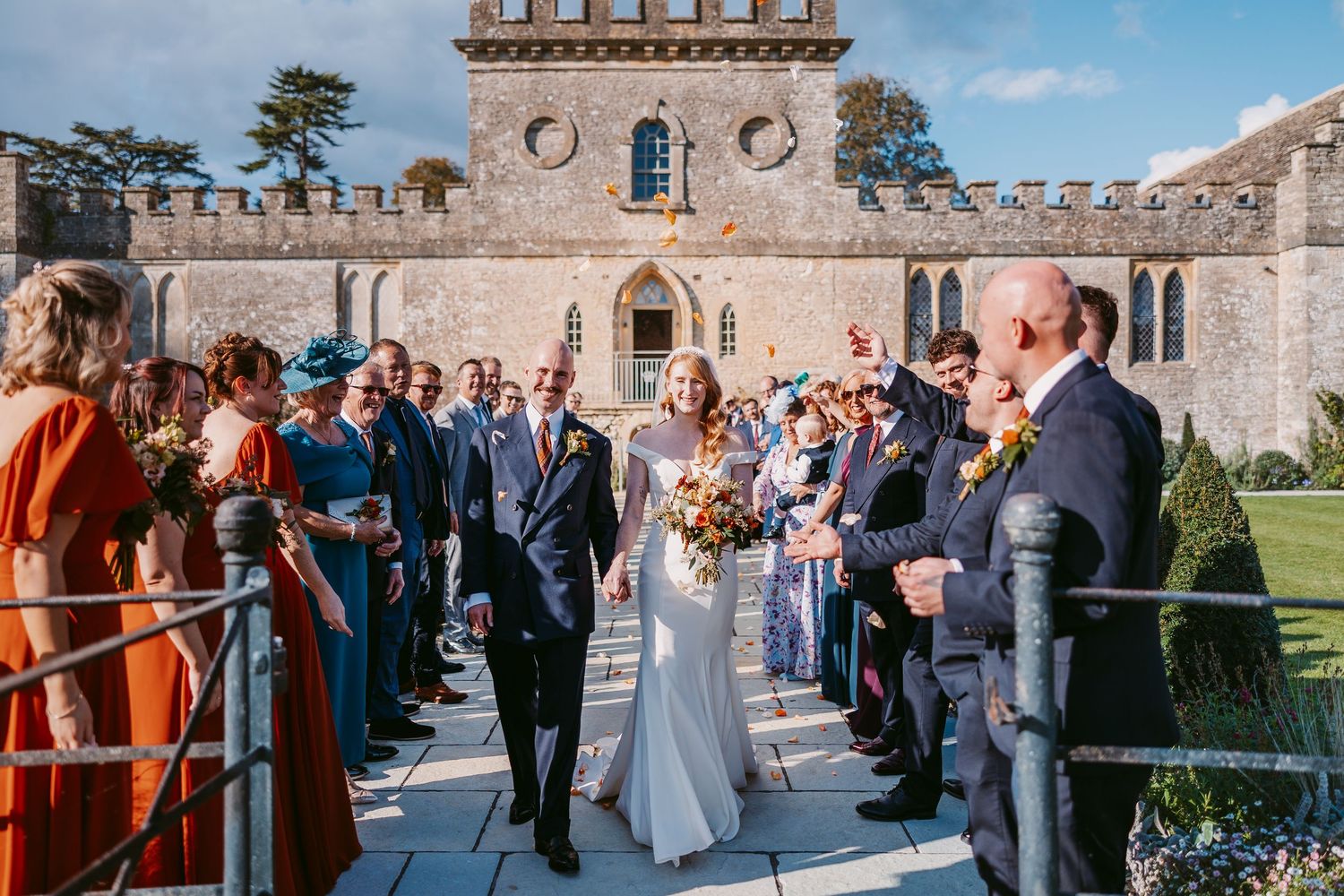 Bride and groom walk hand in hand past cheering wedding guests along a stone path outside a historic castle venue.
