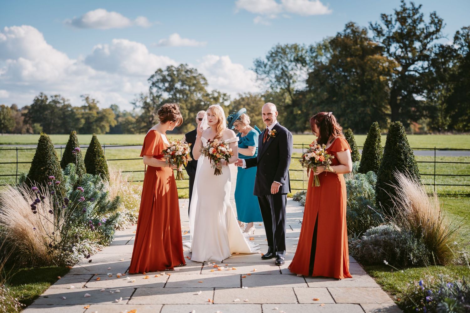 Bride in a white gown and groom in a dark suit stand with bridesmaids in rust and teal dresses on a garden path at an outdoor wedding.