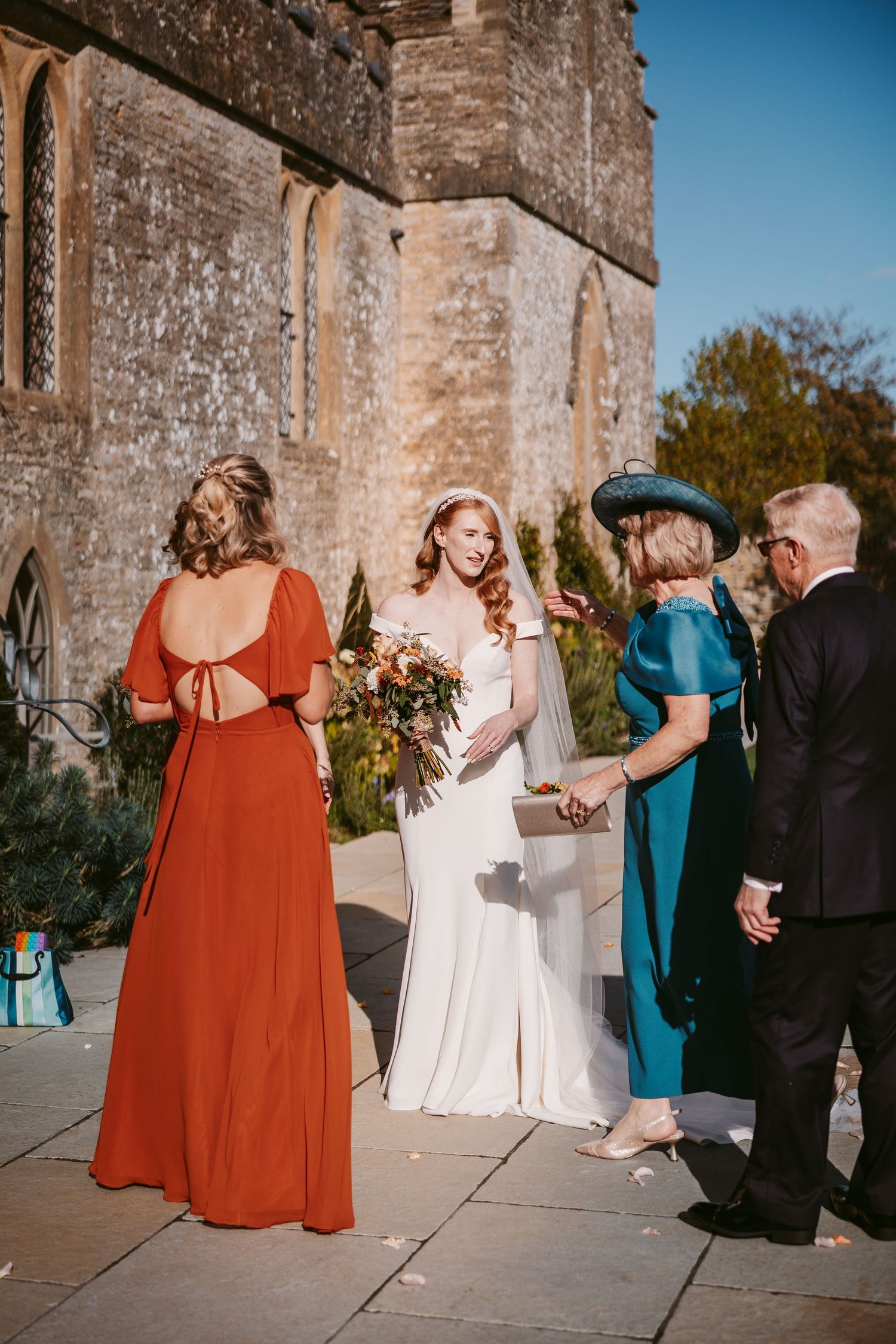 Bride in a white gown and veil smiles at wedding guests in teal and rust dresses outside a historic stone church.