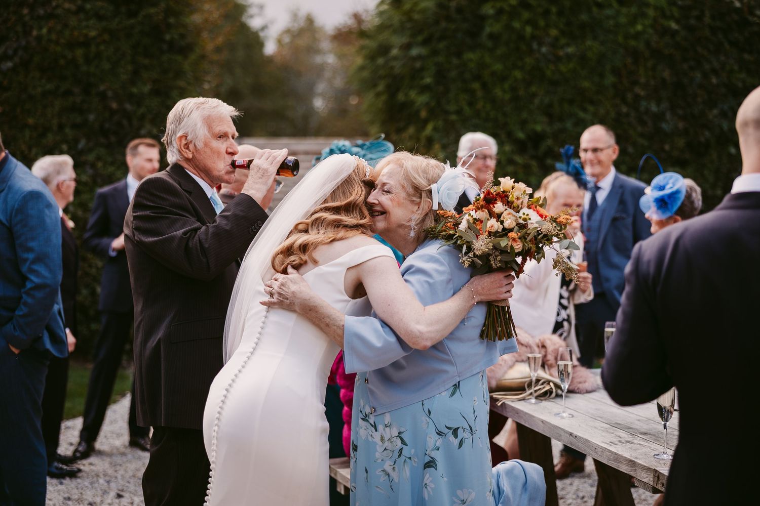 Bride in a white wedding gown hugs an older woman in a blue floral dress while guests watch at an outdoor garden wedding celebration.