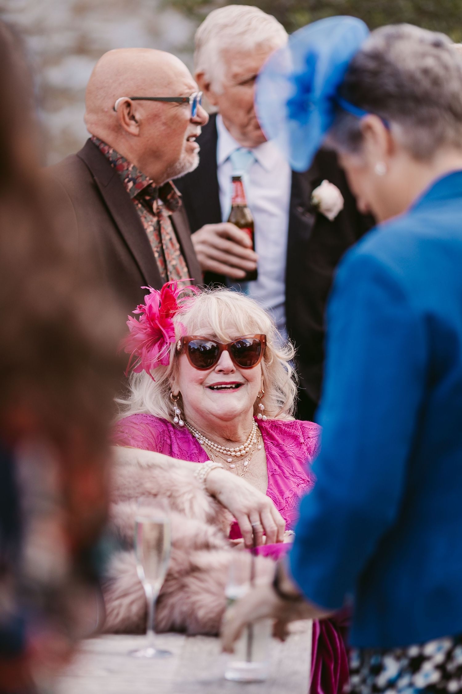 Smiling woman in sunglasses, pink fascinator and pearl necklace sits at a table as guests in formal wedding attire chat behind her.