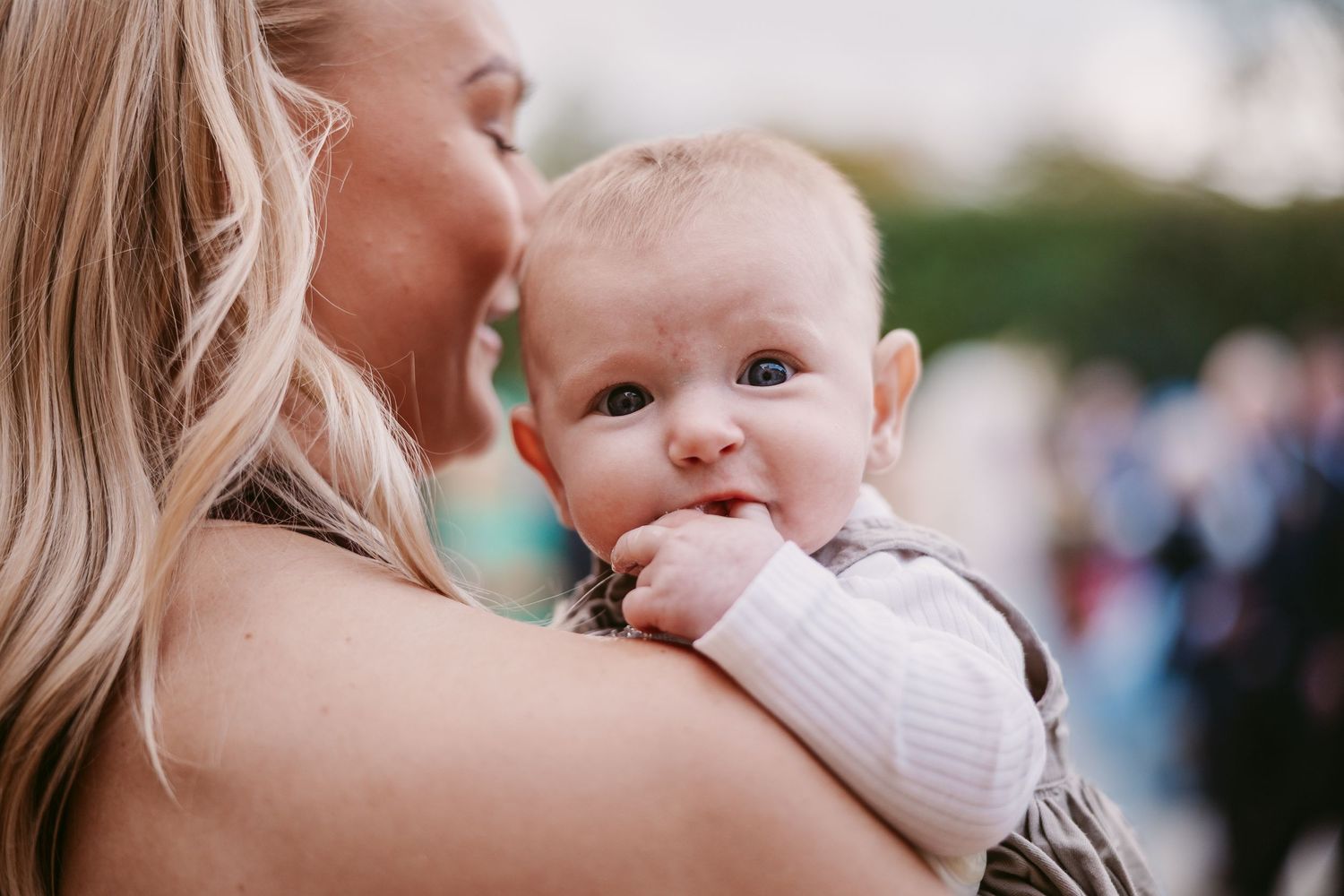 Smiling baby with chubby cheeks chews on a fist while being held over the shoulder of a blonde woman outdoors.