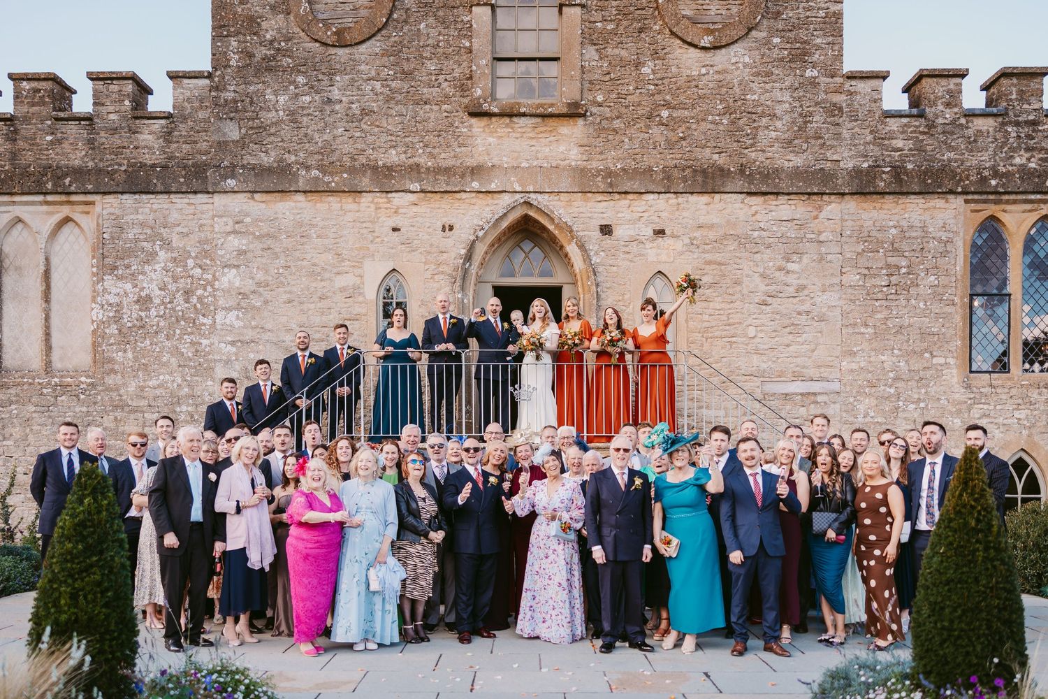Large wedding party in colorful dresses and suits gathered on the steps of a historic stone church on a sunny day.