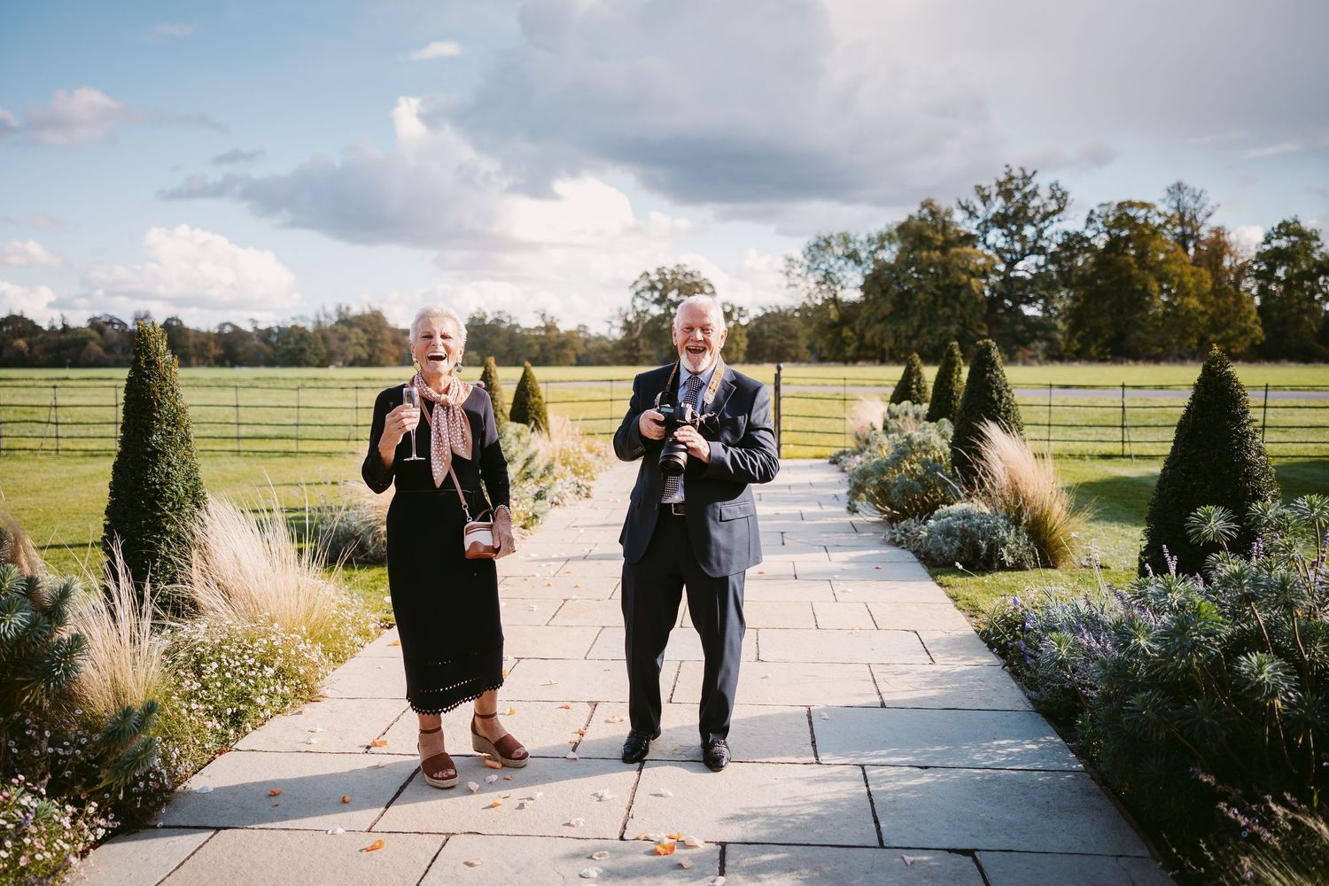 Elderly couple in formal attire stand smiling on a stone garden path lined with shrubs and ornamental grasses, with a grassy field and trees
