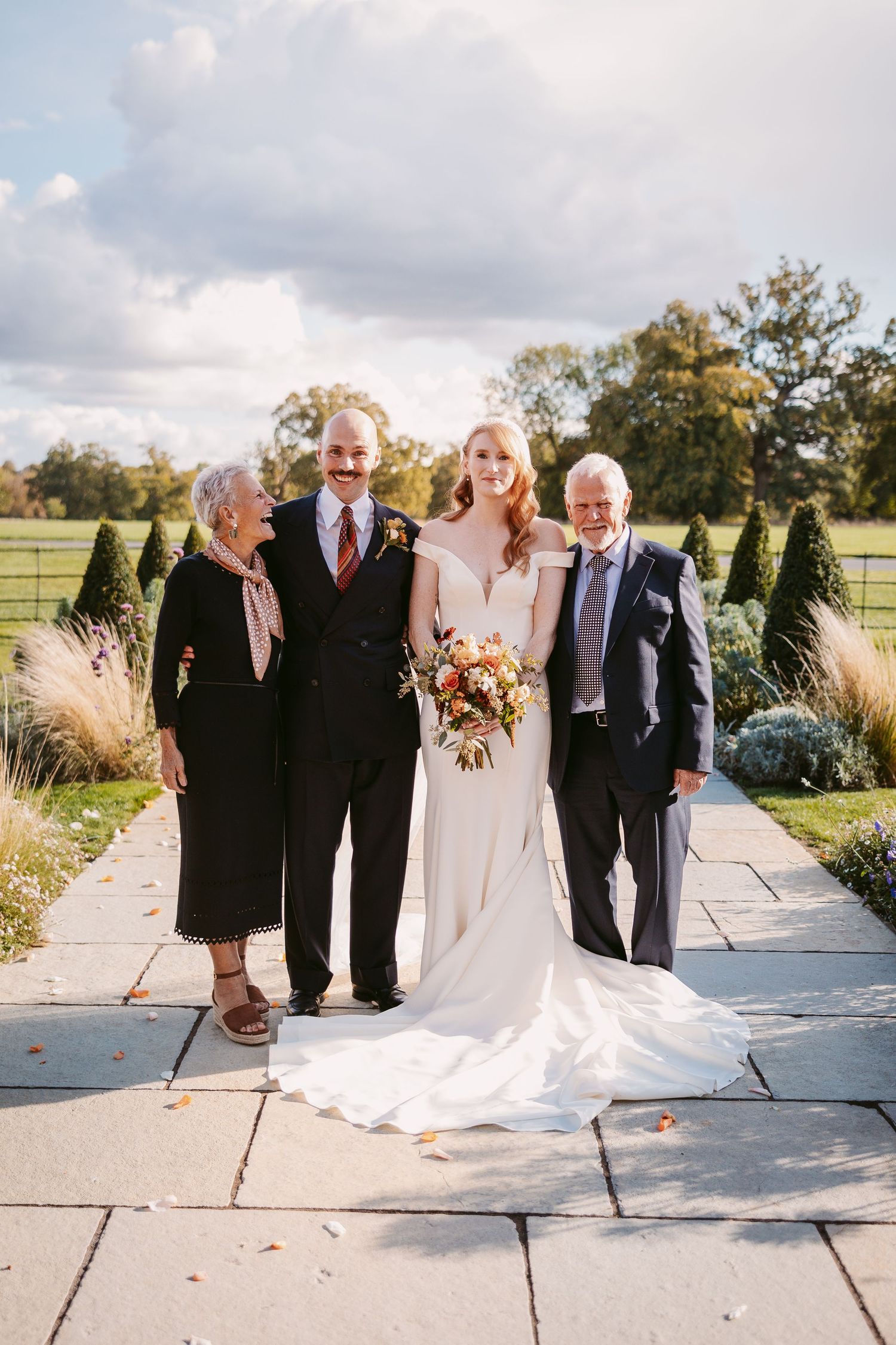 Bride in a white gown holding an autumn bouquet poses outdoors with three family members on a stone patio beside a green lawn.