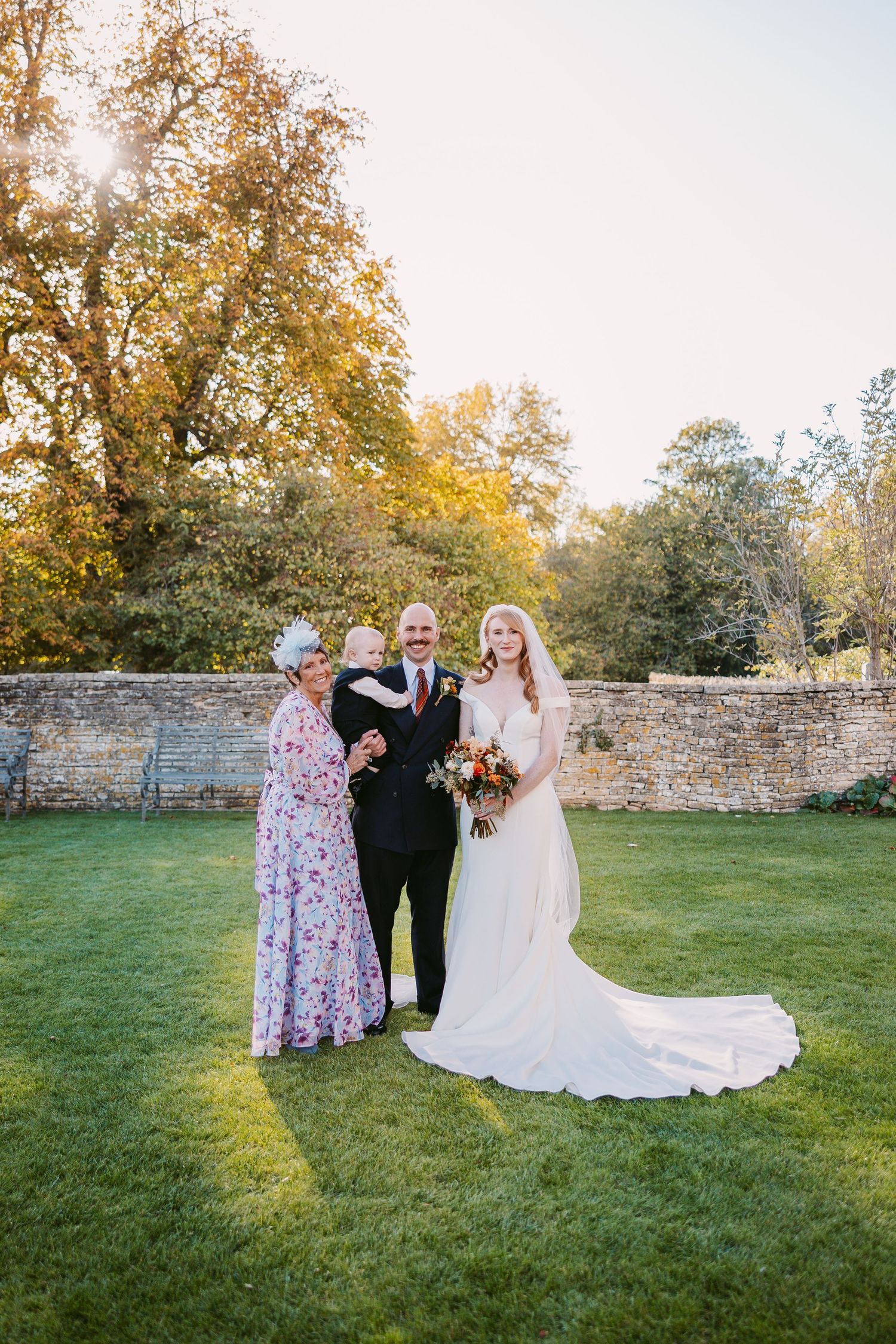 Bride in a white gown poses on a lawn with a groom holding a baby and an older woman in a floral dress beside autumn trees.