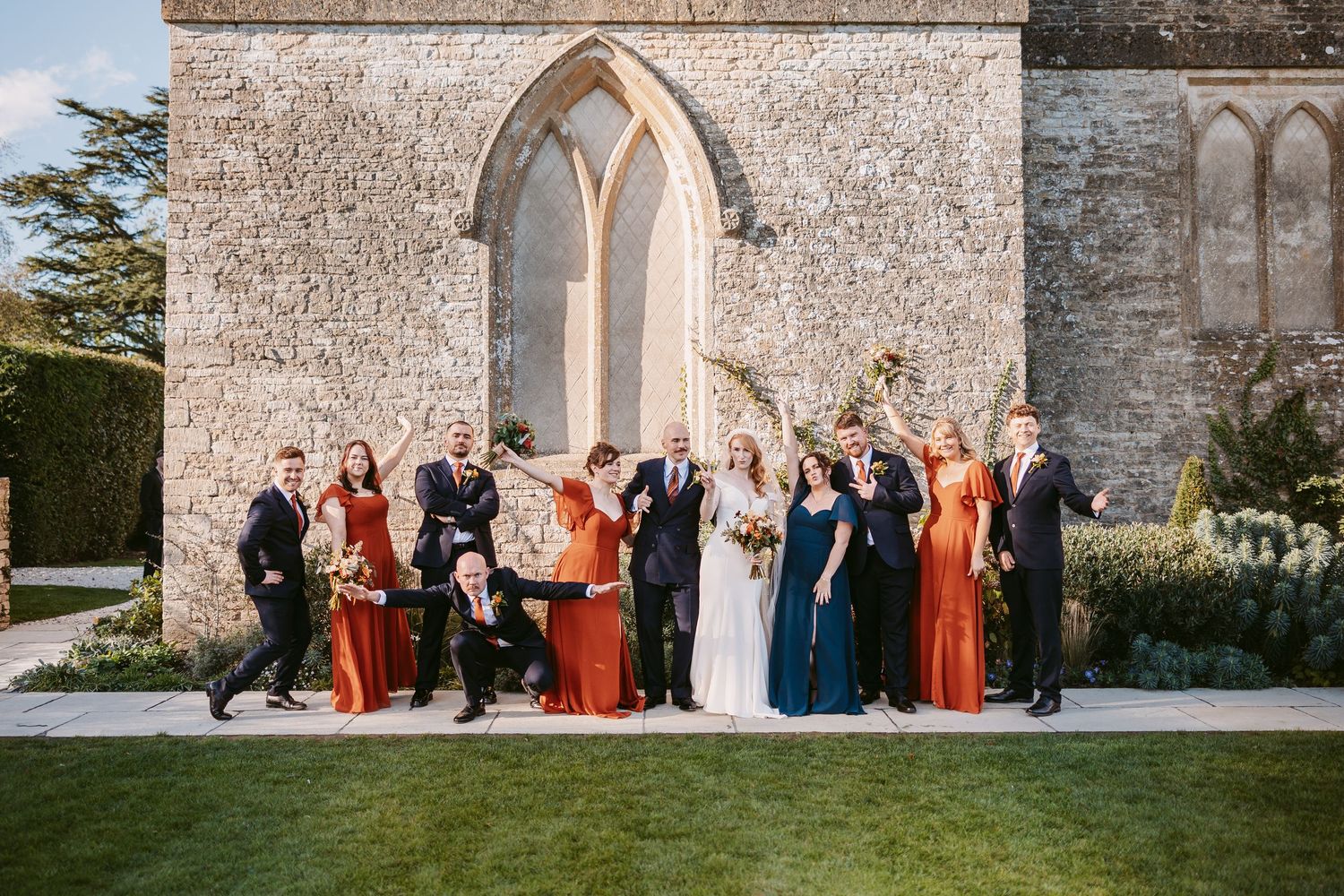 Bride and groom pose with bridesmaids in rust dresses and groomsmen in suits on a lawn before a historic stone church with arched windows.