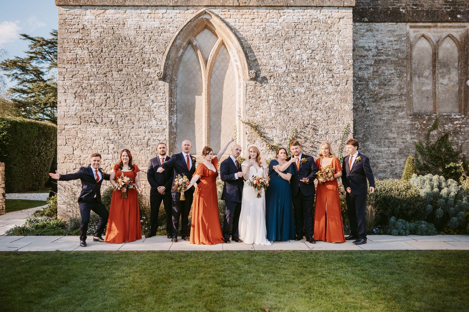 Wedding party in rust and teal dresses and dark suits stands in a line with the bride before a historic stone chapel on a green lawn.