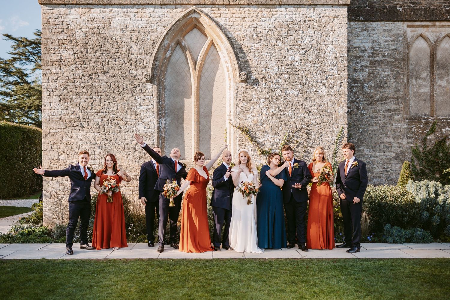 Wedding party in rust and teal dresses and dark suits cheer beside a bride in white before a historic stone church with arched windows.