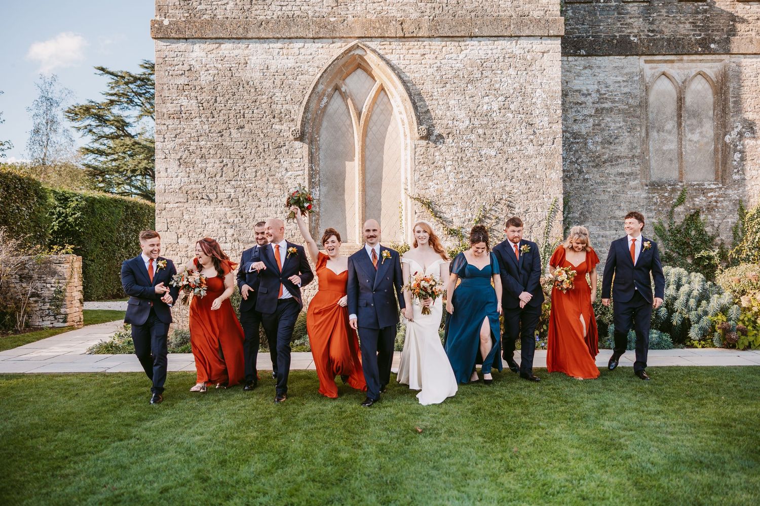 Bride and groom walk across a lawn with bridesmaids in rust orange dresses and groomsmen in suits in front of a stone church.